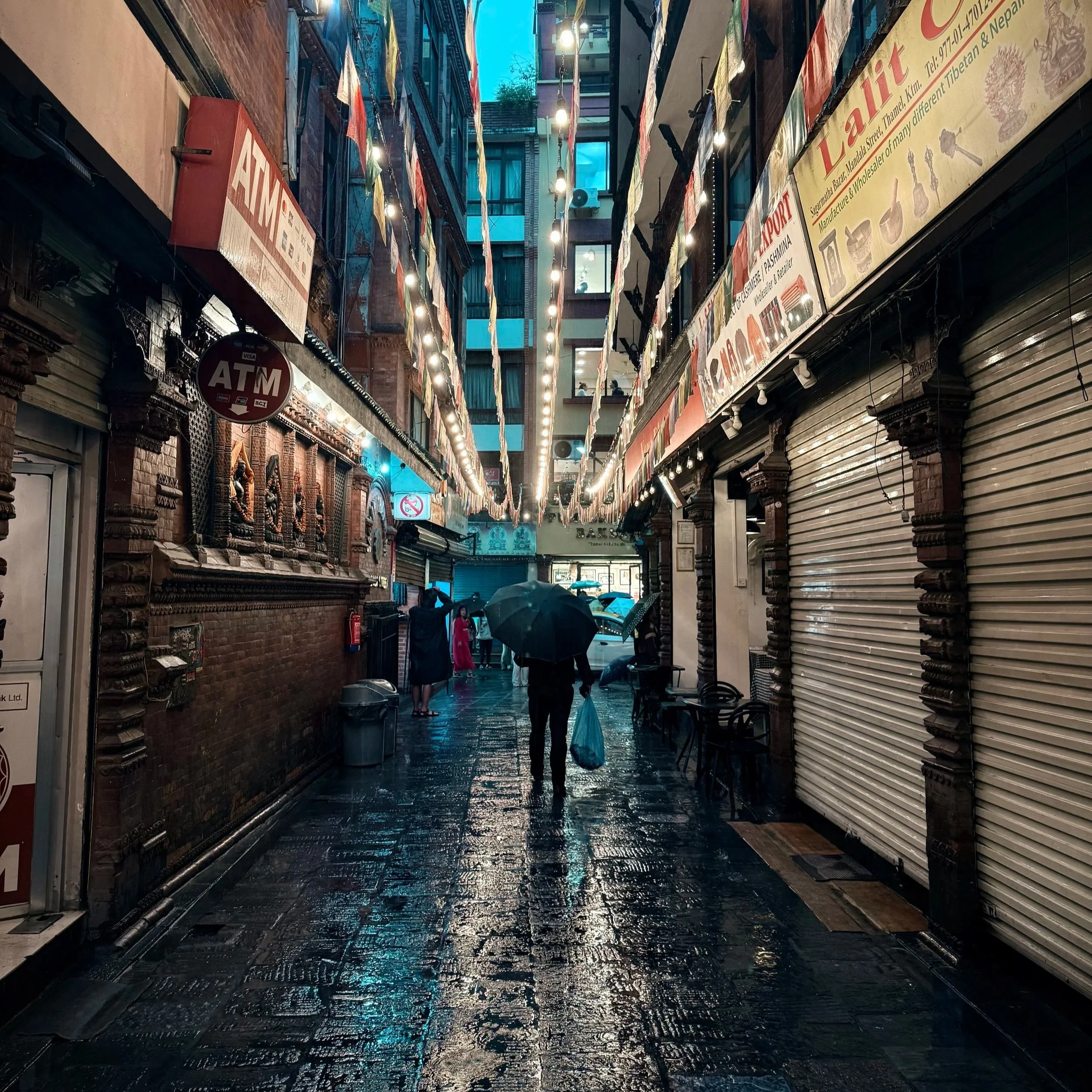 Lone figure with umbrella walking through a rain-slicked alley strung with colorful prayer flags, Thamel district, Kathmandu, Nepal