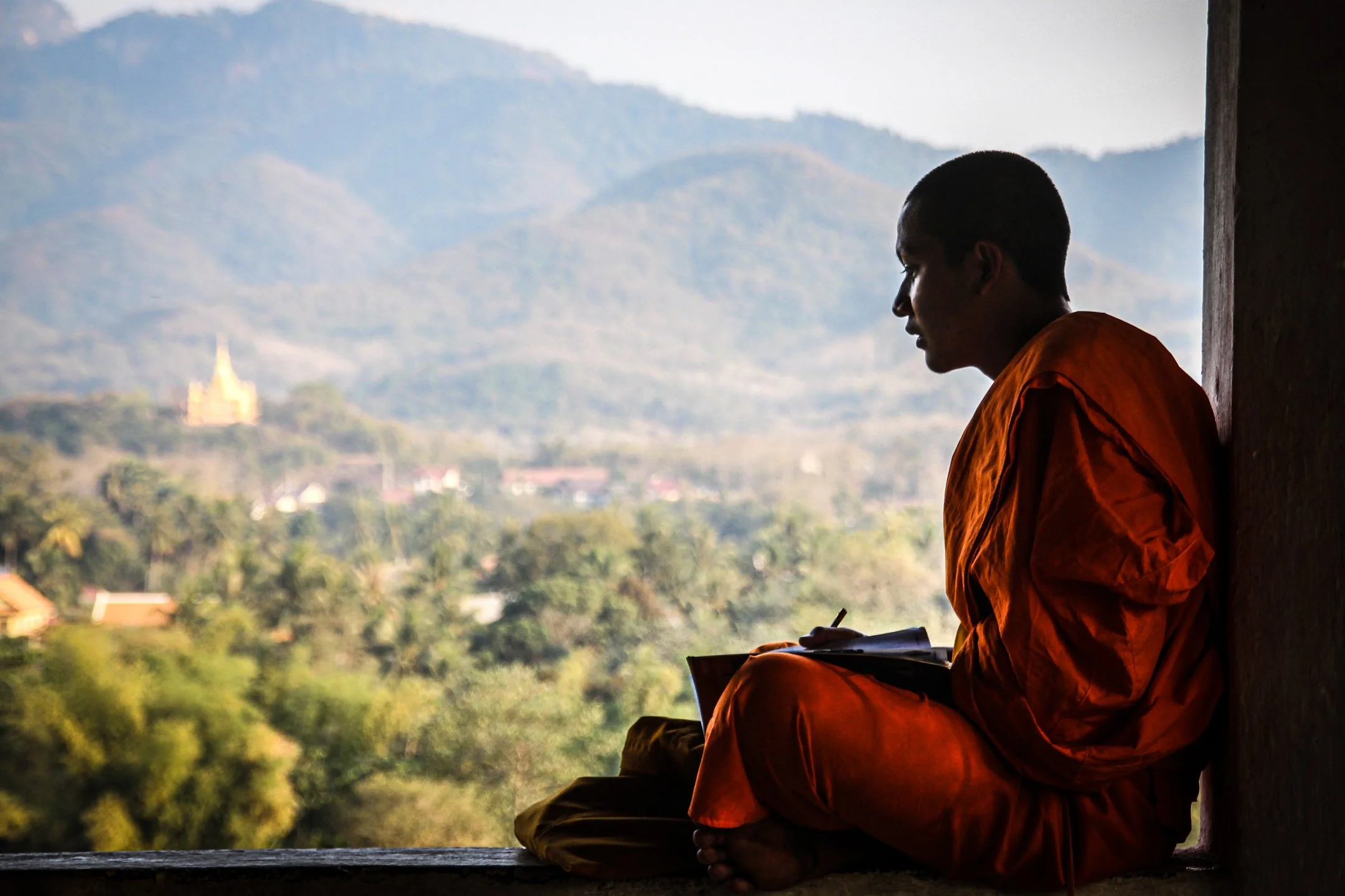 Buddhist monk seated in a window overlooking the misty green hills and temples of Luang Prabang, Laos