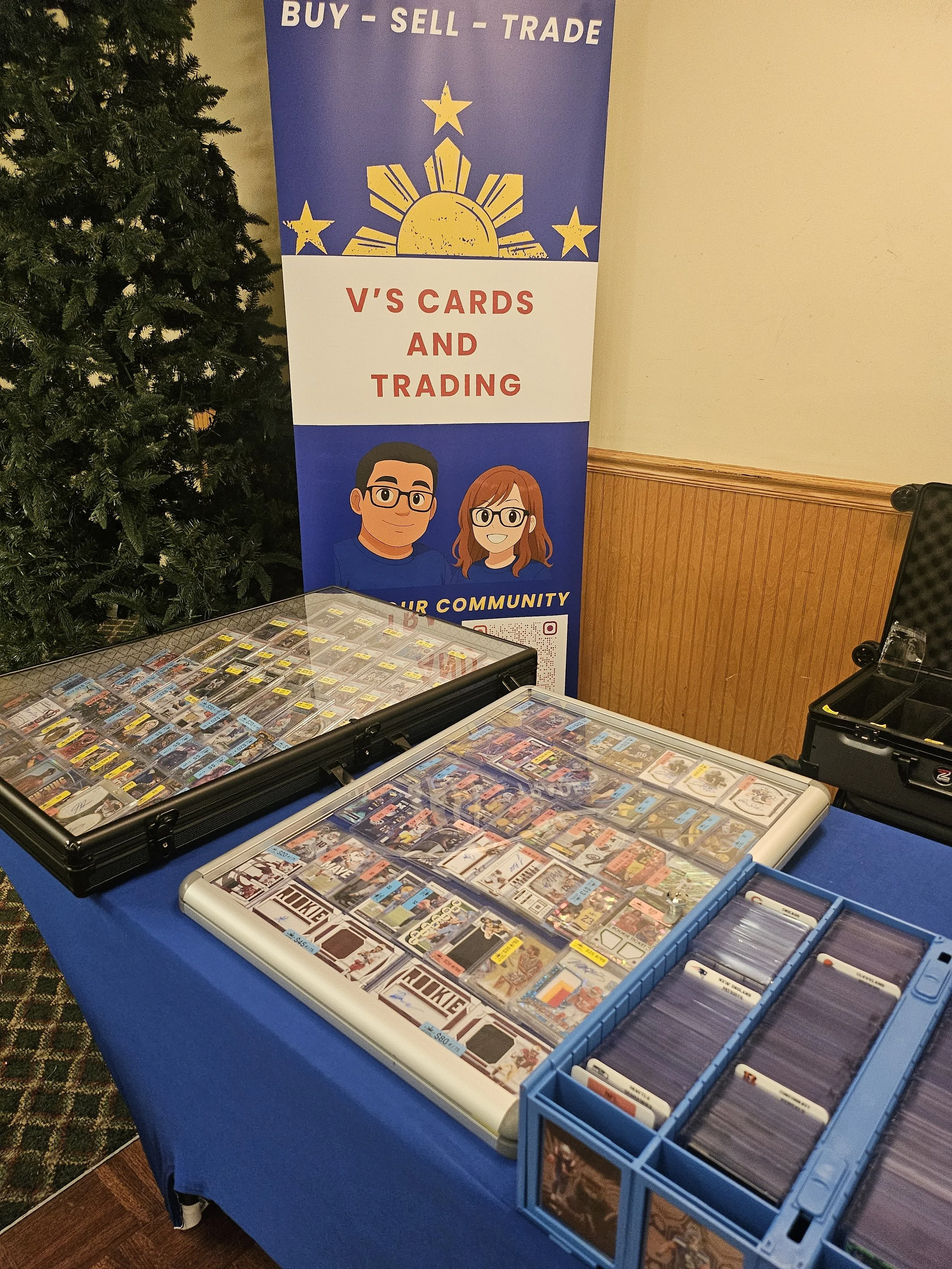 Table displaying trading cards in cases, with a banner in the background promoting buy, sell, trade activities from V's Cards and Trading.