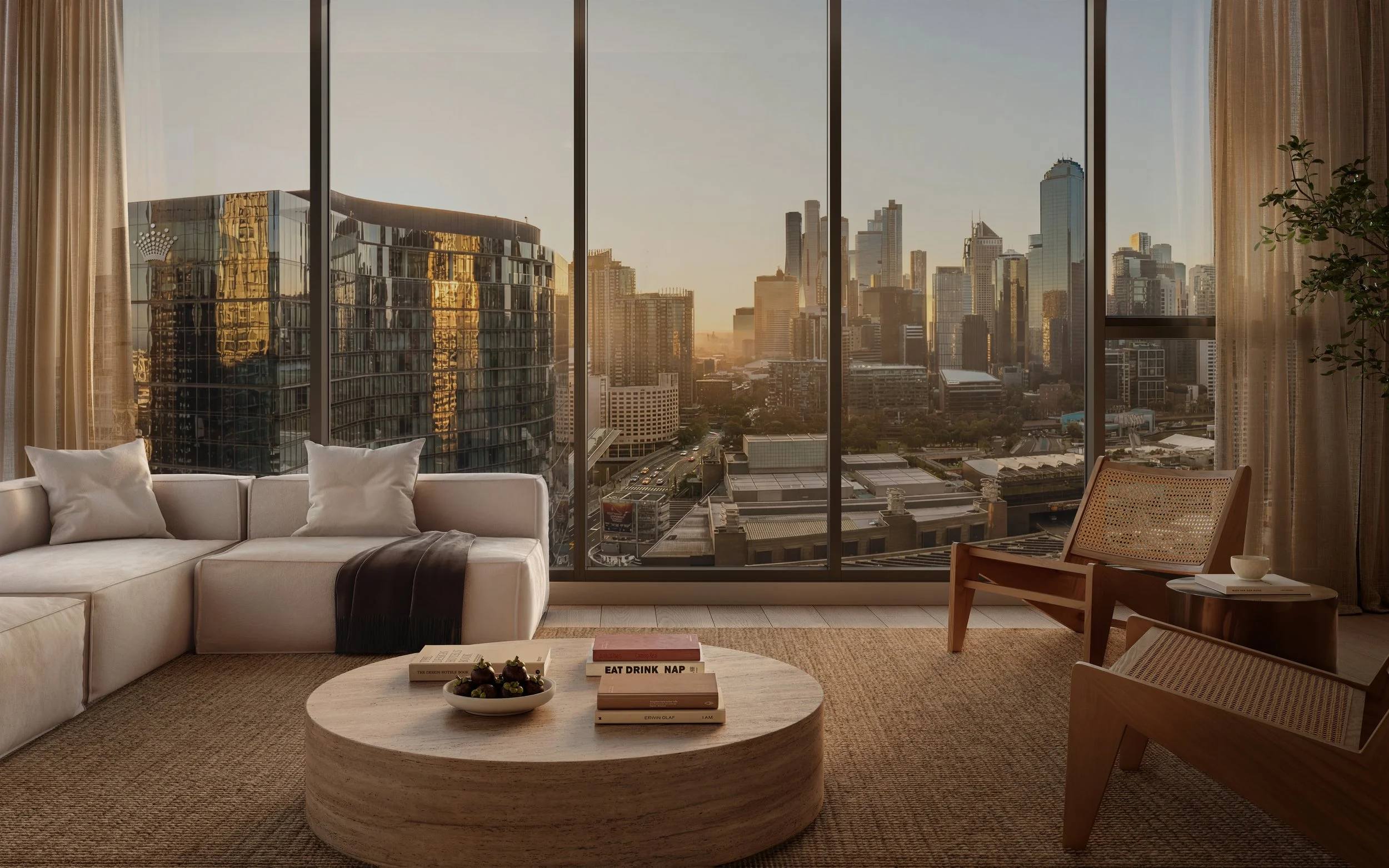 Living room with large window showing a city skyline at sunset. Features a white sofa with pillows, a round coffee table with books and a fruit bowl, wooden chairs, and beige curtains.
