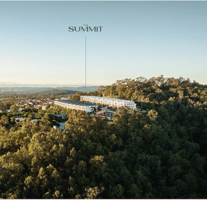 Aerial view of a hilltop development with white buildings, surrounded by dense green trees, under a clear sky, with a label pointing to the location called "The Summit."