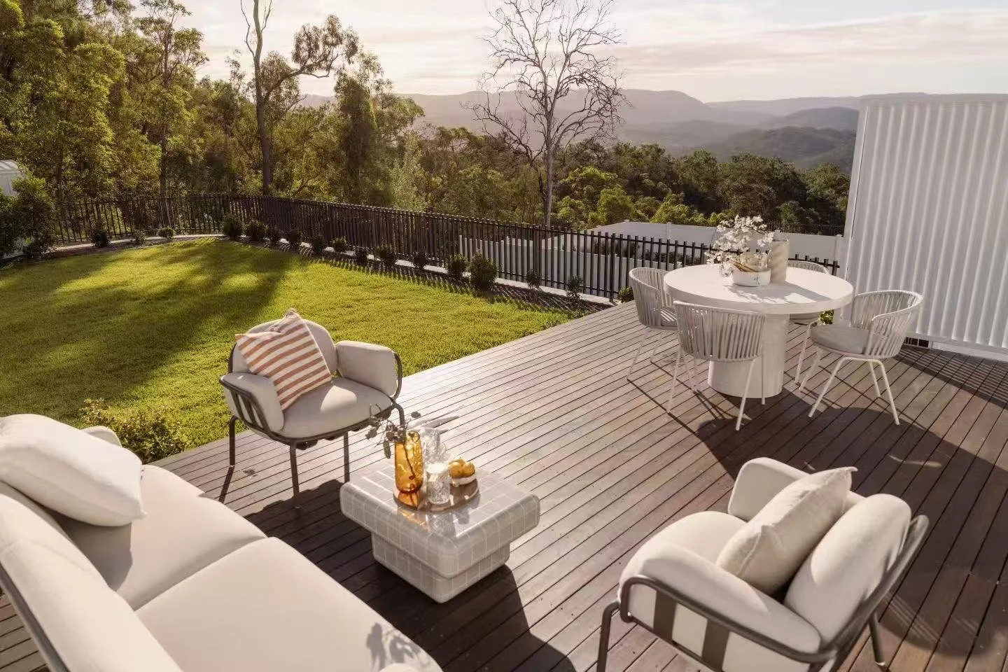 An outdoor wooden deck with white furniture overlooking a green yard and distant hills. The deck has a sofa, armchairs, a small table with drinks, and a round dining table with chairs.
