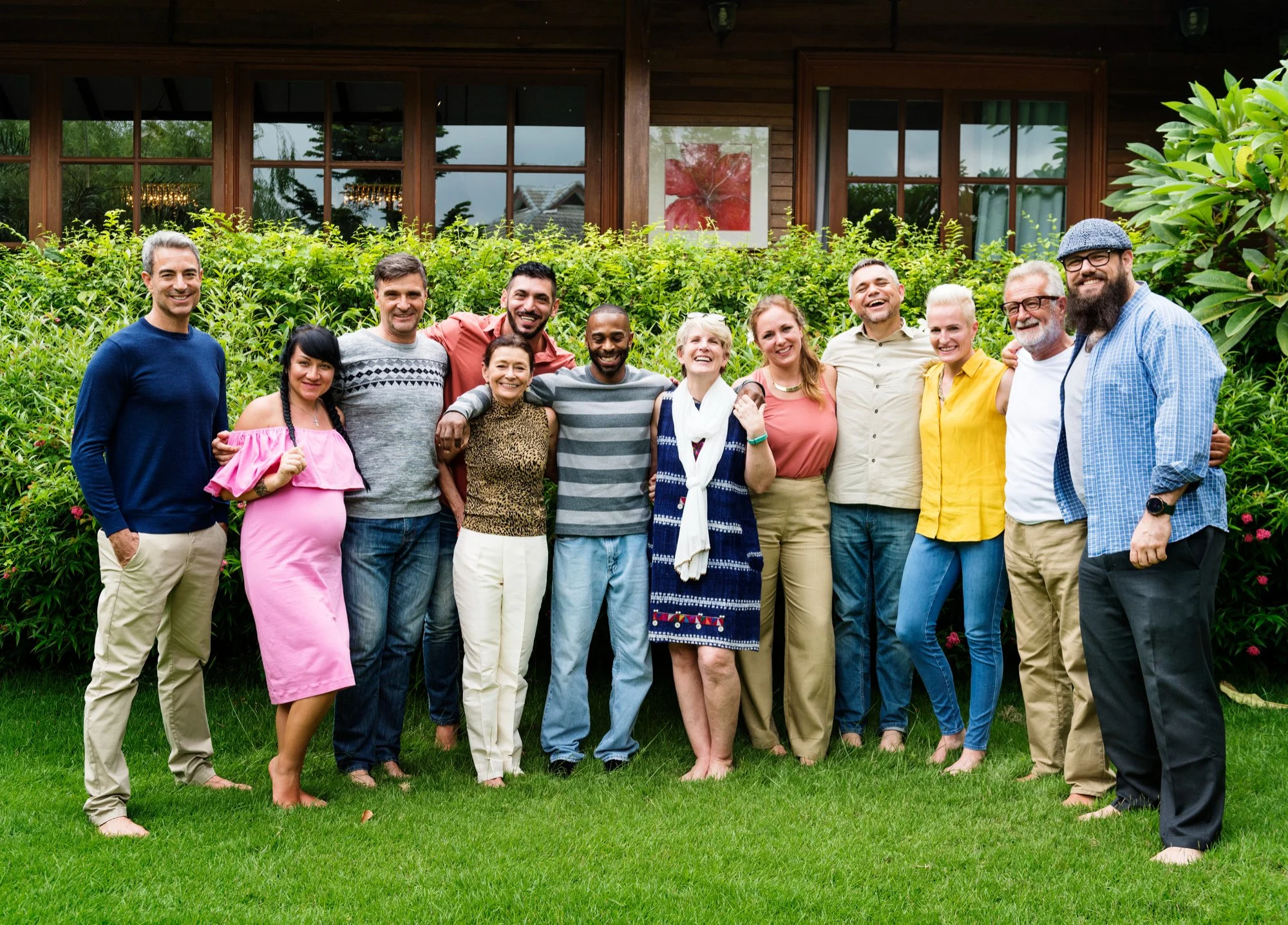 A group of 13 diverse people standing outdoors on a lawn, smiling and posing with arms around each other, in front of a house with large windows and green bushes.