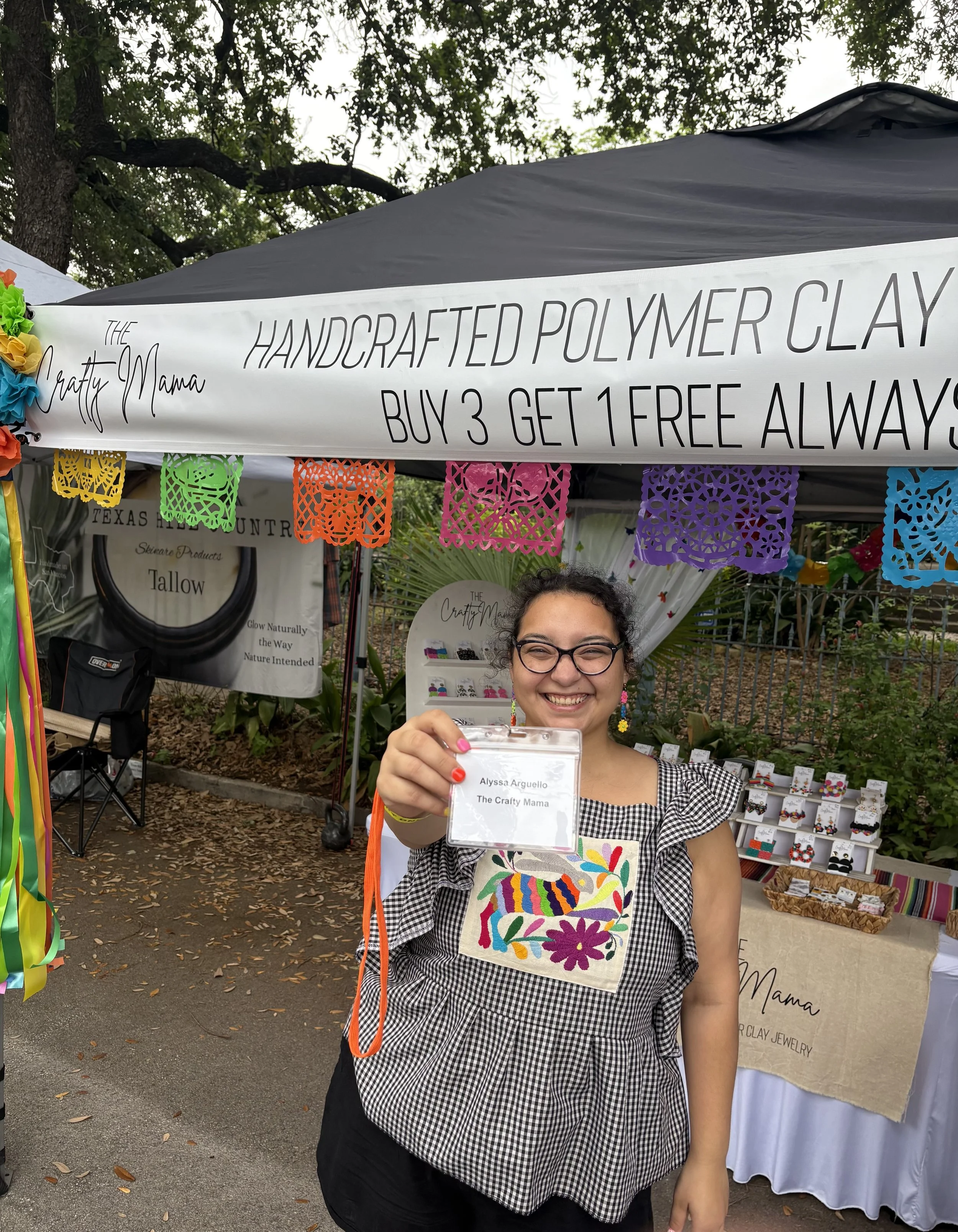 A smiling woman with glasses holding a clear plastic badge with her name, 'Alyssa Arguello,' and her business, 'The Crafty Mama.' She is standing in front of a booth at an outdoor craft fair with colorful papel picado banners, jewelry displays, and a cafe tent in the background.