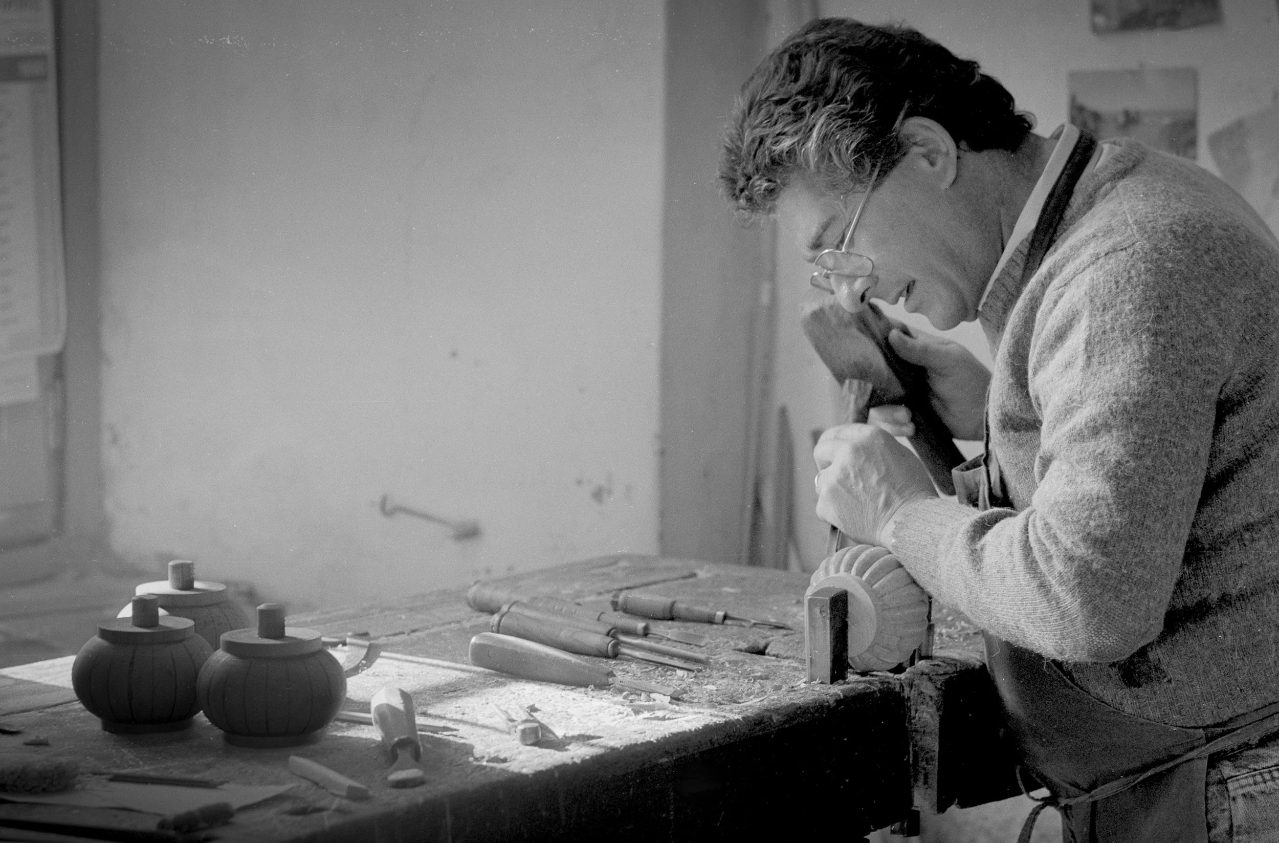 A artisan in Florence, Italy, with glasses, wearing a sweater, is working on a wooden ornament at a table in a studio, surrounded by completed pieces.