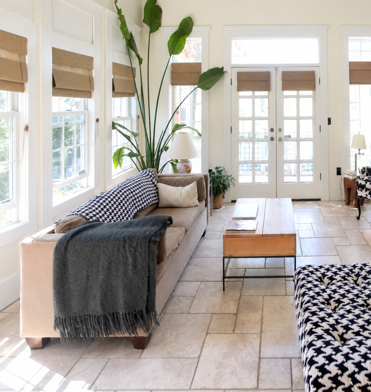 Living room with beige sofa, black and white patterned throw blanket, white pillows, large green plant, wooden coffee table, floor lamp, and French doors with glass panels.
