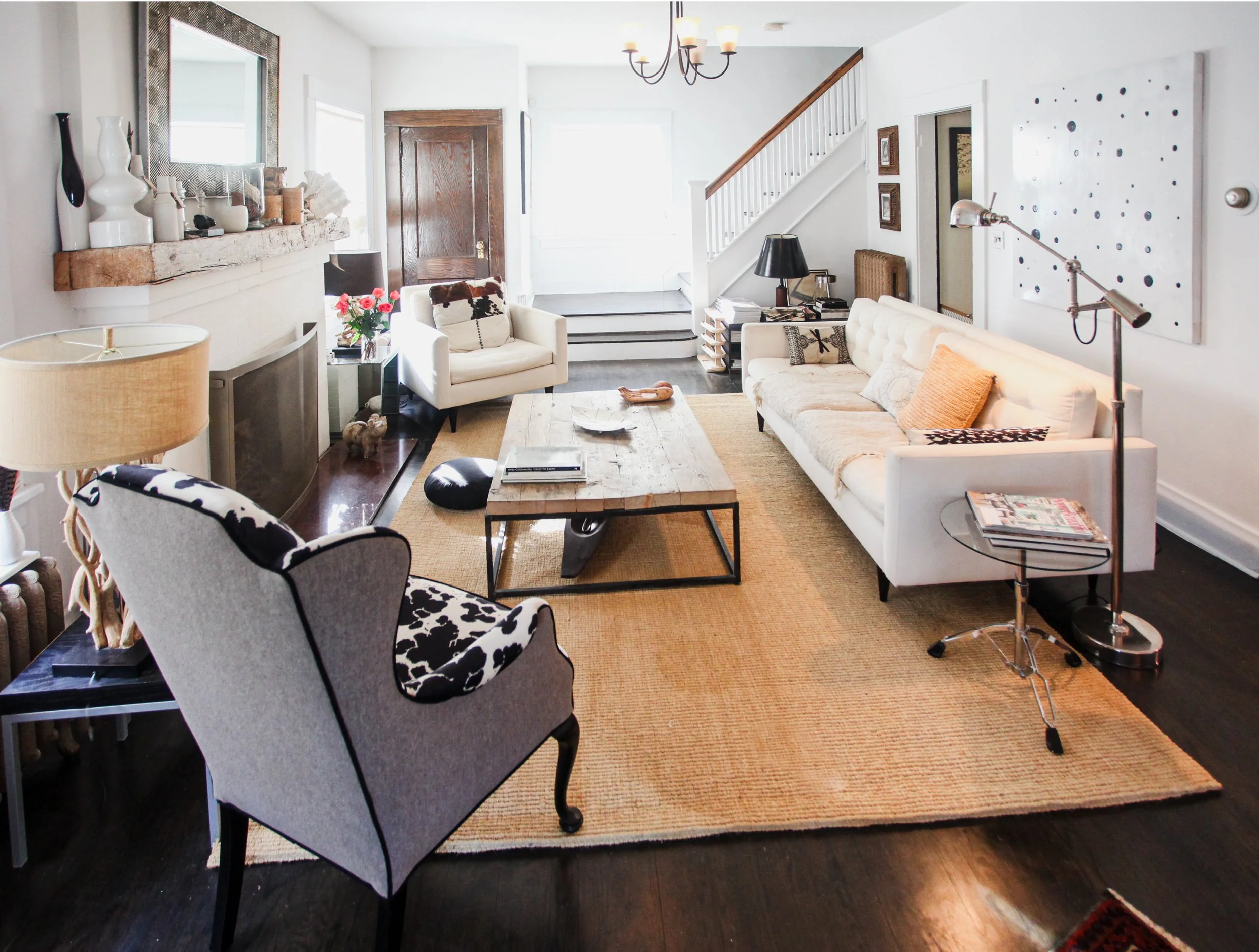 Living room with white walls, a beige sofa, a white armchair, a coffee table, and a chandelier. There are decorative vases on a mantel and a large artwork on the wall. Stairs lead to another level, and a writing desk with a desk lamp is visible.