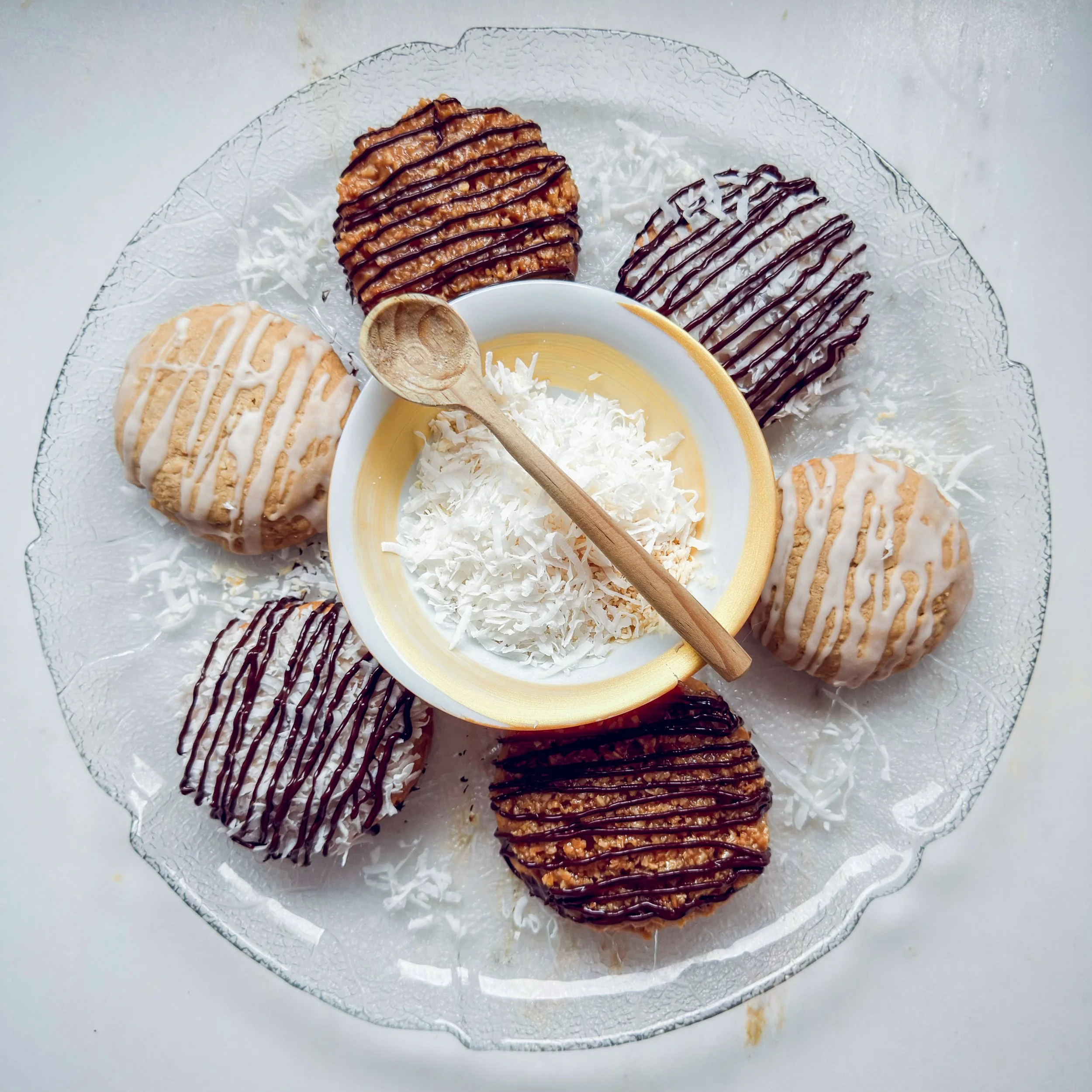 Assorted cookies on a glass plate with drizzled chocolate and white icing, served with a small bowl of yellow custard or pudding topped with shredded coconut, a wooden spoon.