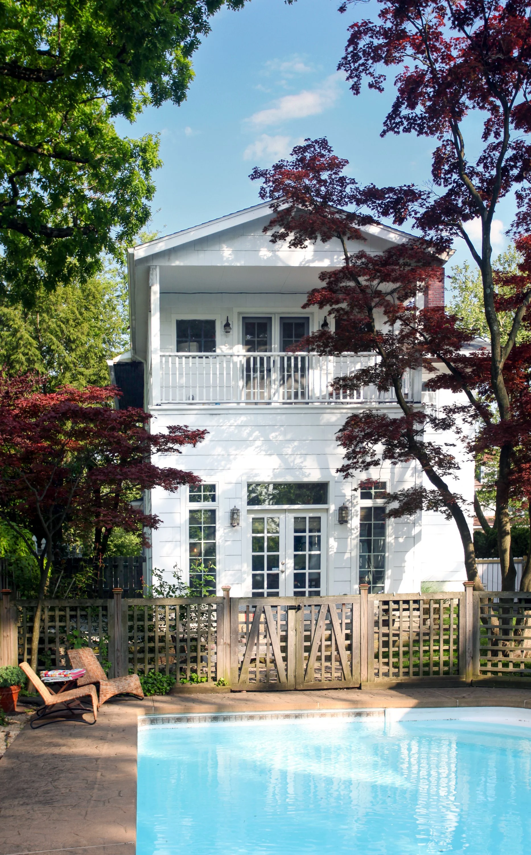 A backyard scene featuring a swimming pool, a wooden fence, a patio chair, and a white house with a second-story balcony surrounded by green and red trees.