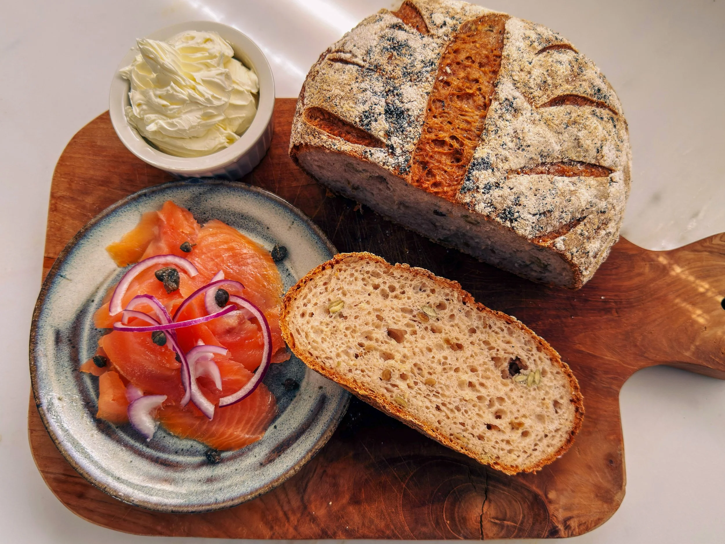 A loaf of rustic bread with a dusting of flour, alongside a small bowl of whipped butter, slices of smoked salmon with red onion and capers, and slices of toasted bread, all arranged on a wooden serving board.