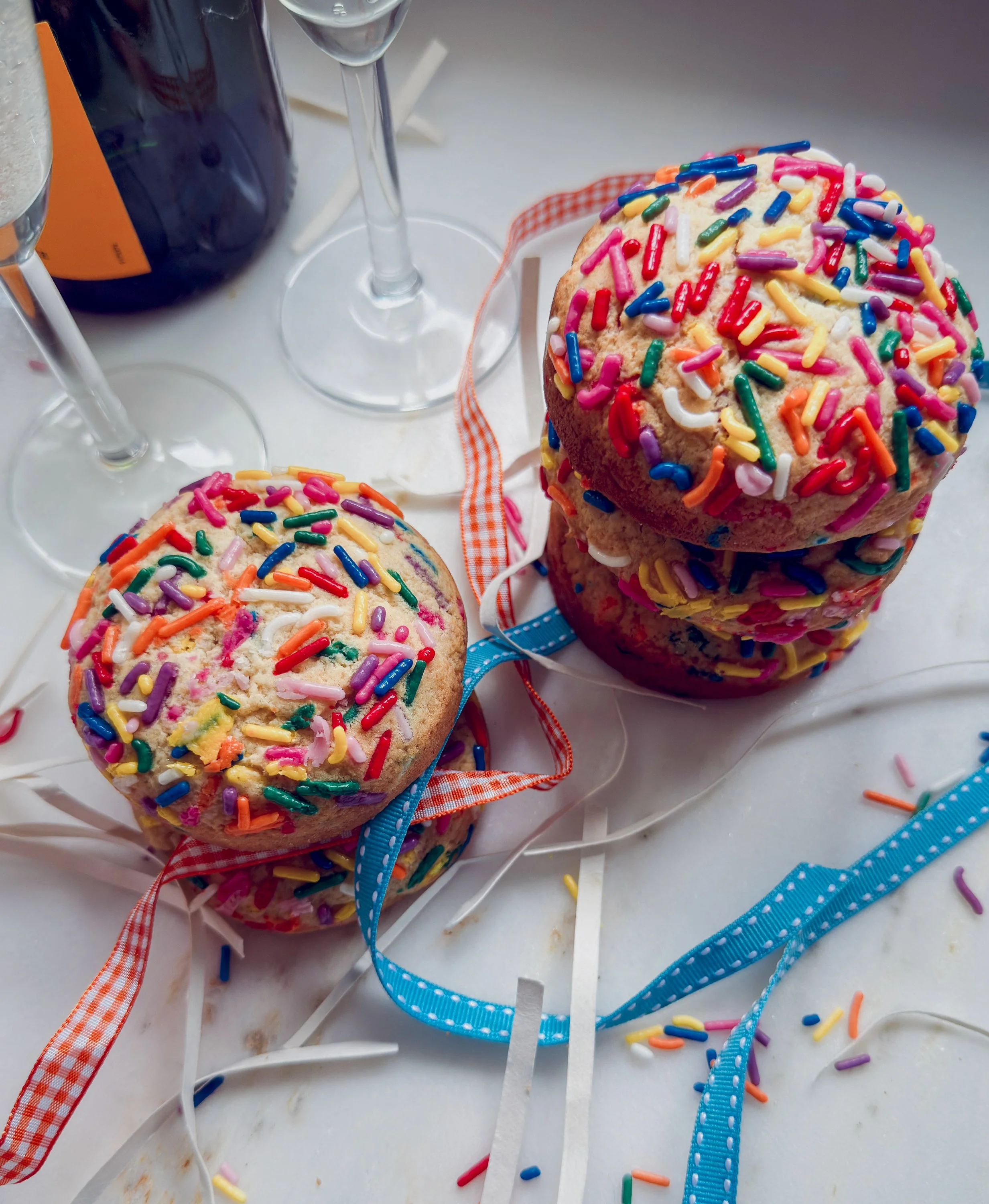 Close-up photo of colorful birthday cupcakes with sprinkles on top, ribbons around the base, and some scattered sprinkles and ribbons around on a white surface.