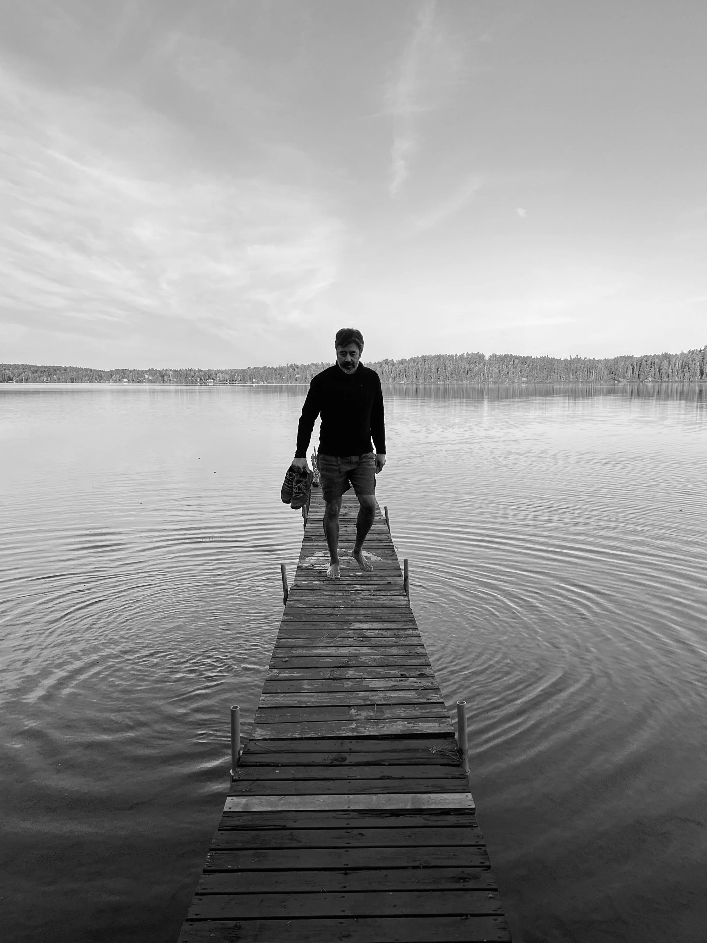 A man is walking barefoot on a small wooden pier over a calm lake in remote Canada, holding shoes in one hand, with trees and sky in the distance.