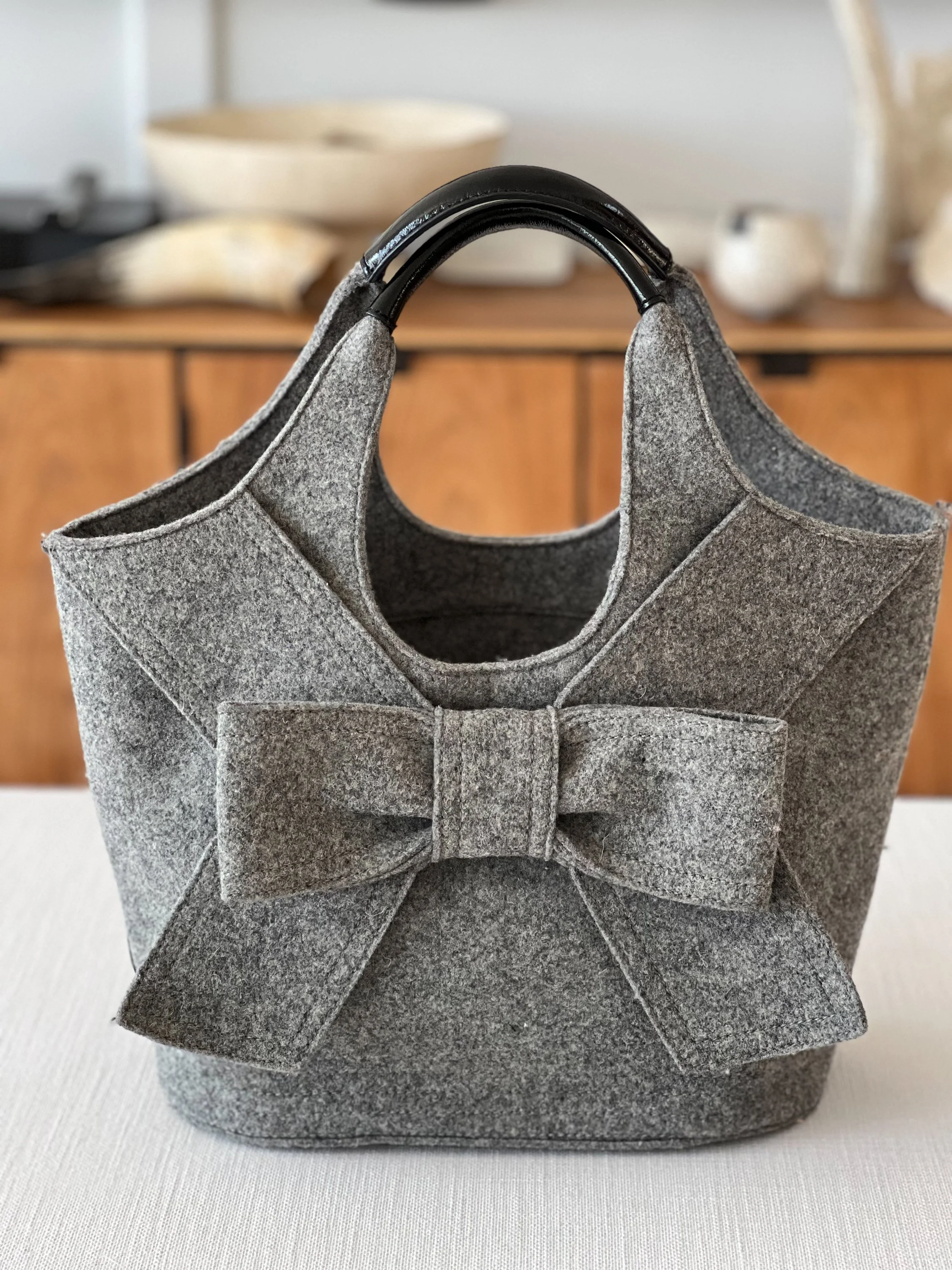 Gray felt Kate Spade tote bag with a bow detail on the front, hanging on a chair with a blurred background of kitchen bowls and pottery.