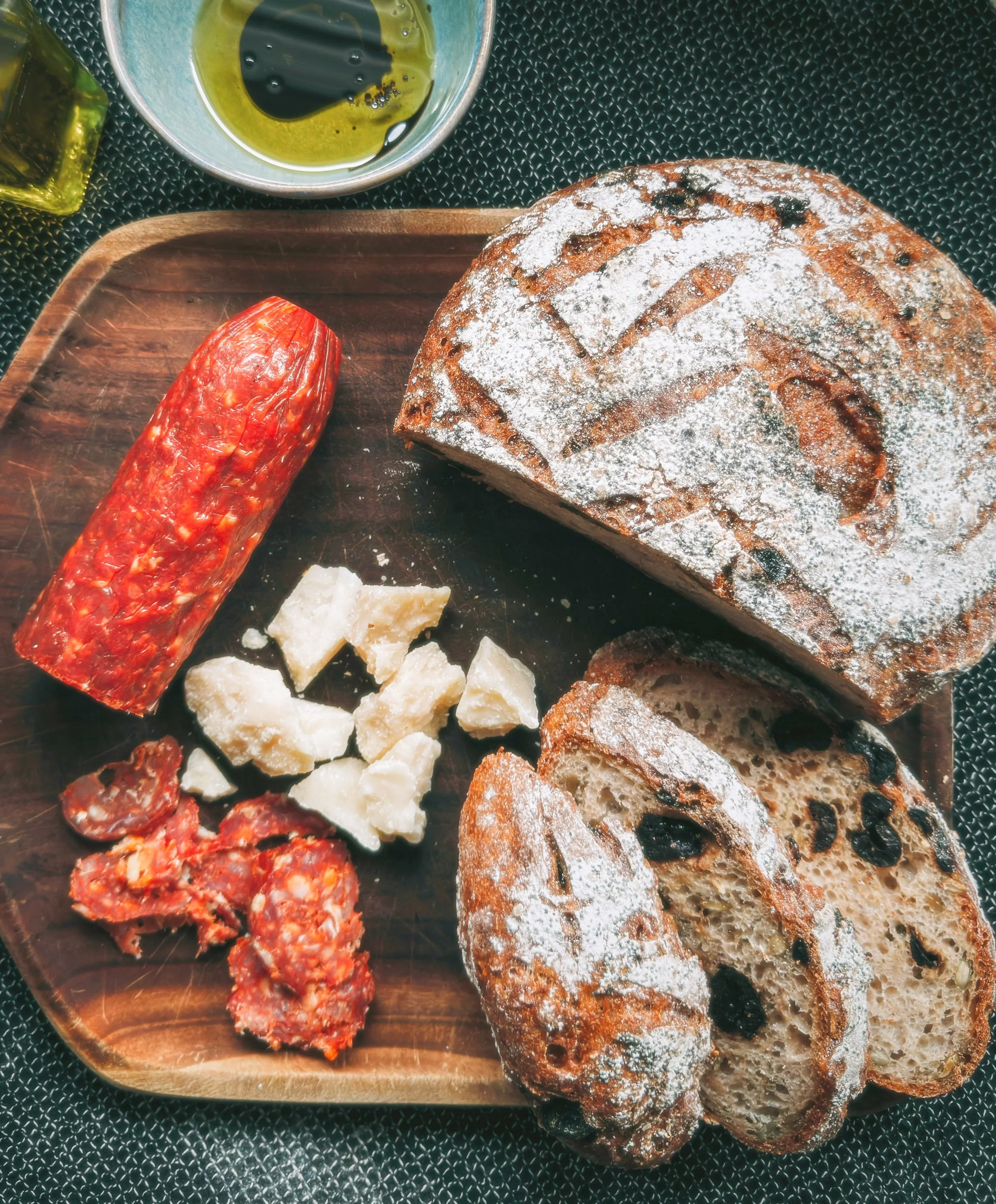 A wooden tray with slices of crusty bread dusted with flour, a slice of chorizo sausage, and chunks of blue cheese. In the background, a metal dish with olive oil and balsamic vinegar, and a small bottle of olive oil.