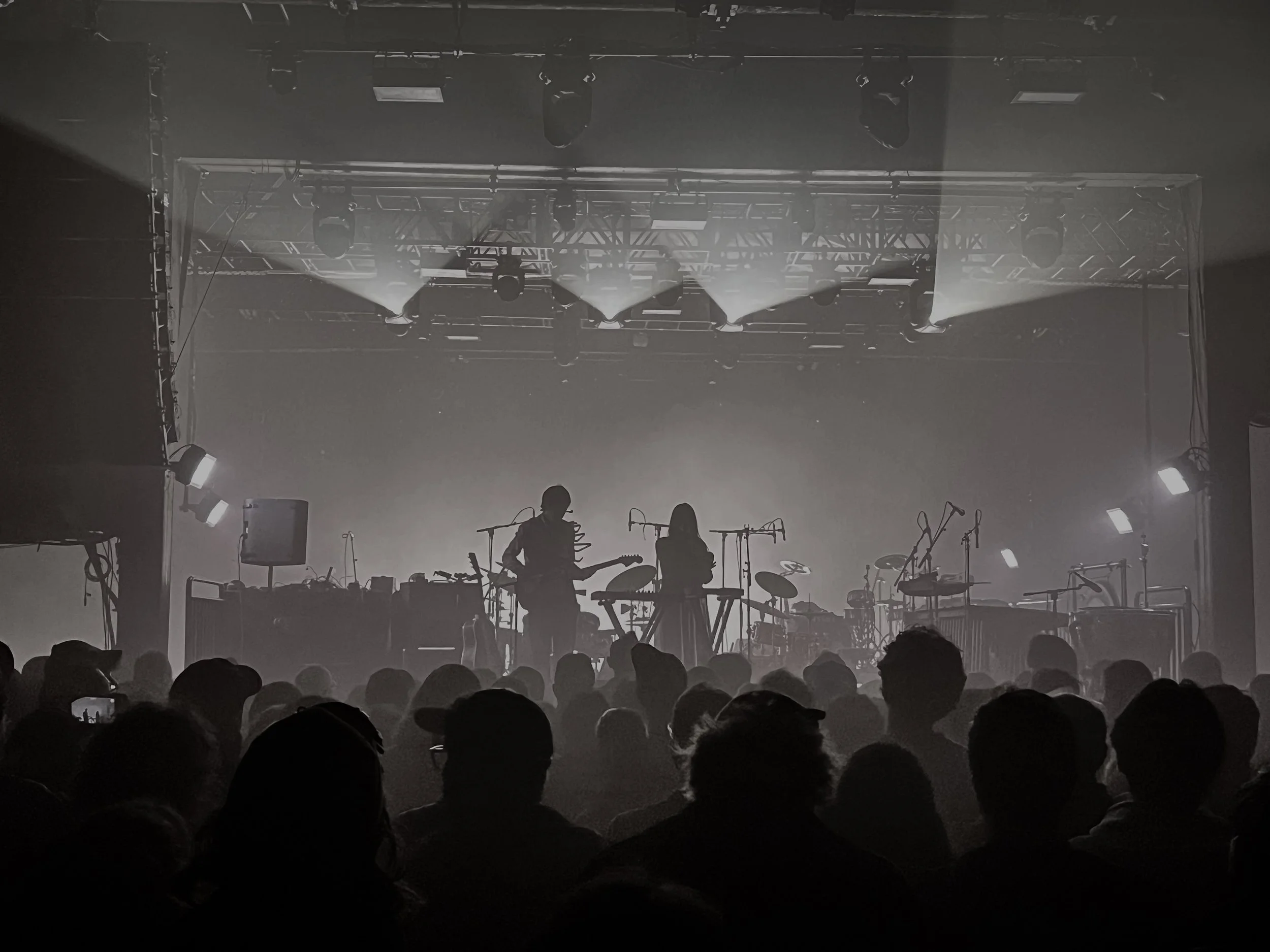 A concert stage with multiple spotlights and fog, featuring silhouetted musicians and a large audience in front.