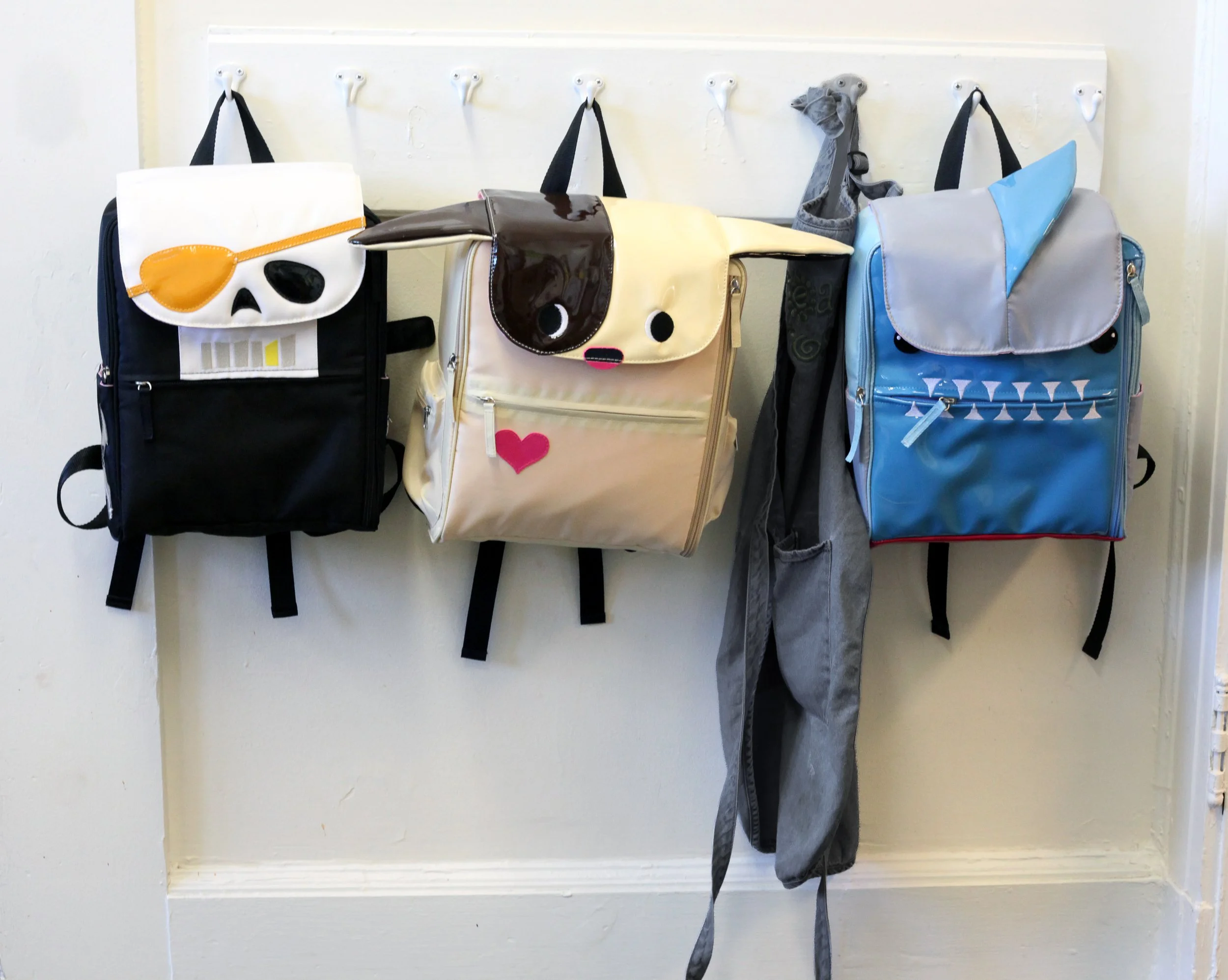 Three children's backpacks by Warm Milk, Inc. hanging on hooks on a white wall. The backpacks have animal face designs resembling a panda, a dog, and an elephant.