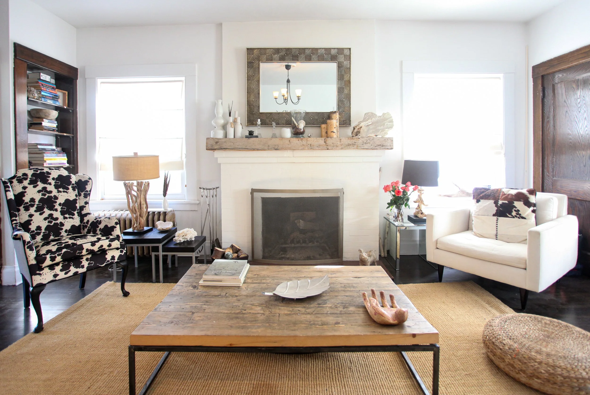 Living room with a wooden coffee table, a beige rug, a cow print armchair, a white sofa with black and white pillows, a fireplace decorated with vases and shells, a mirror above the fireplace, and two windows letting in natural light.
