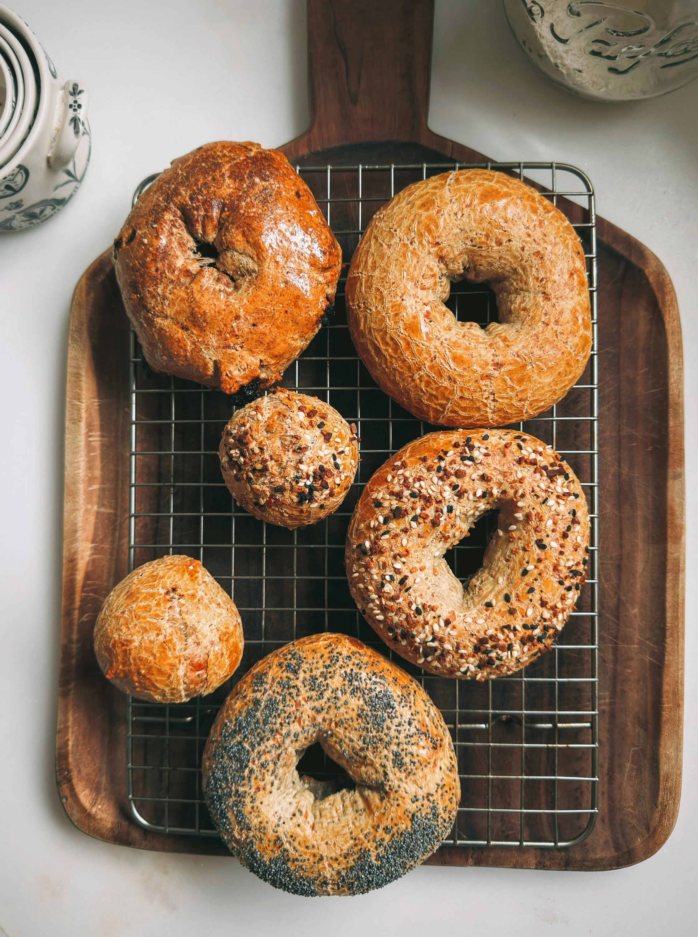 assorted bagels on a wire rack, including plain, everything, poppy seed, and sesame seed varieties, on a wooden tray with a white background.