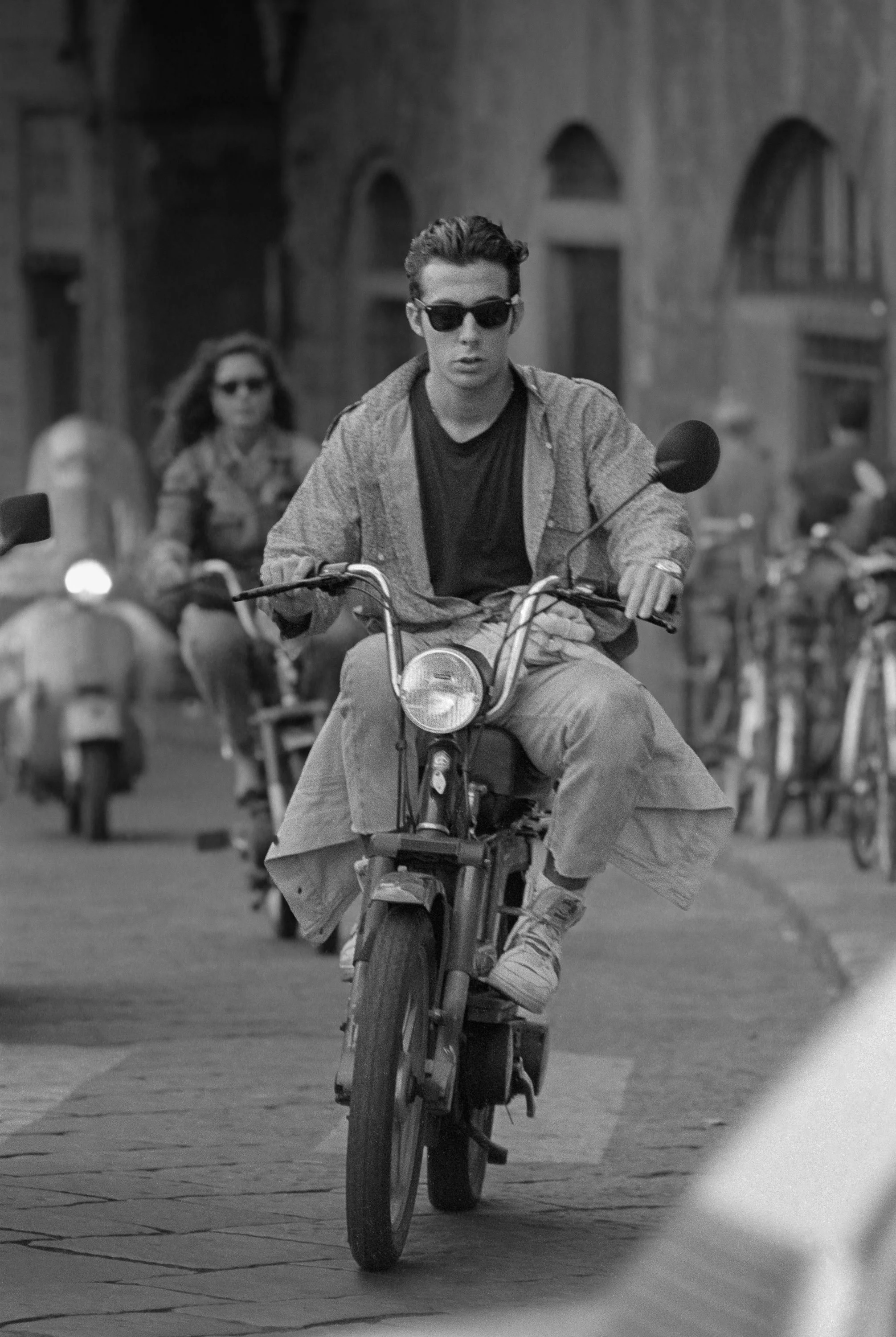 A young man riding a bicycle in Florence, Italy, wearing sunglasses, a hoodie, and a t-shirt, with a woman on a bicycle in the background.