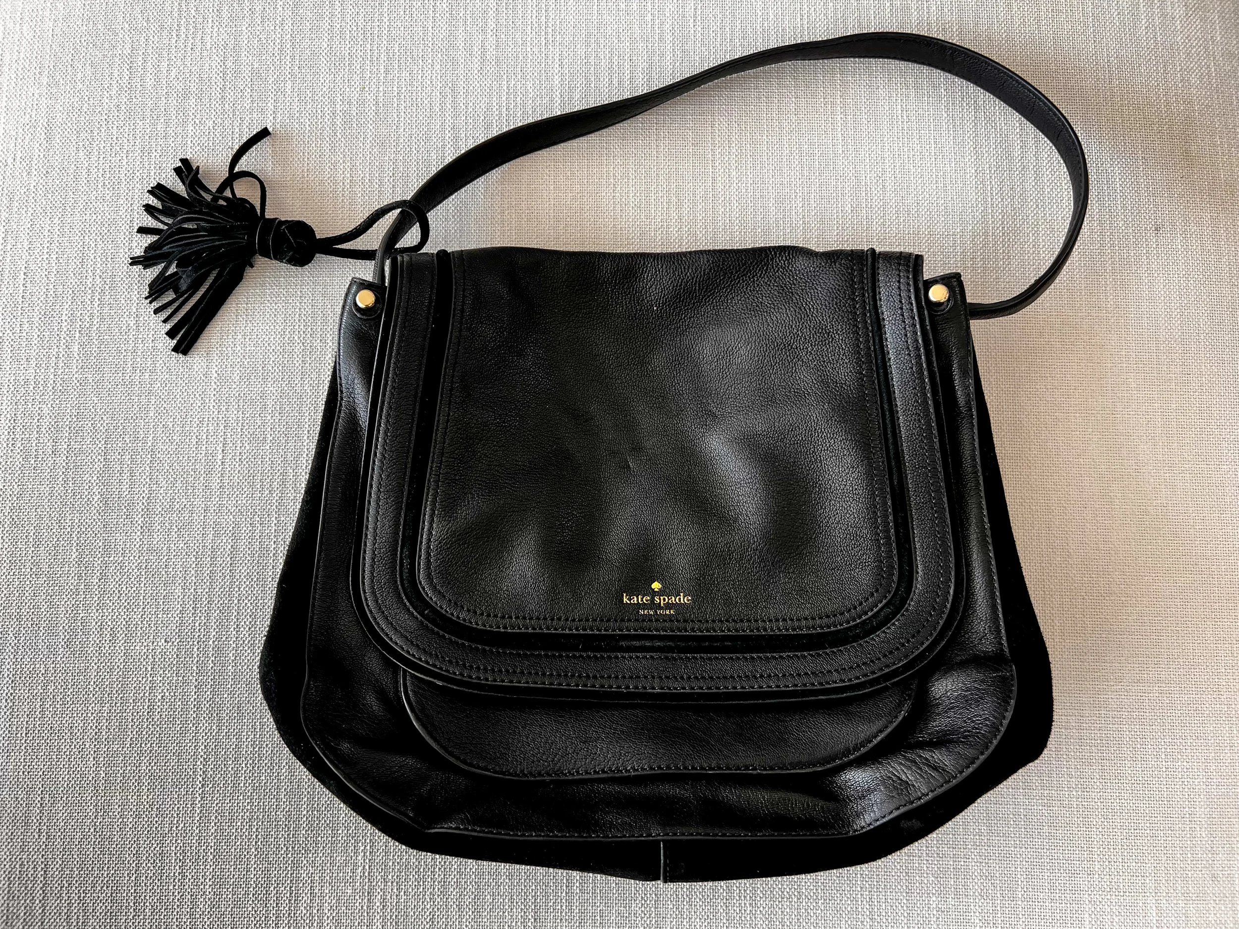Black leather Kate Spade shoulder bag with fringe detail and gold accents on a textured gray background.