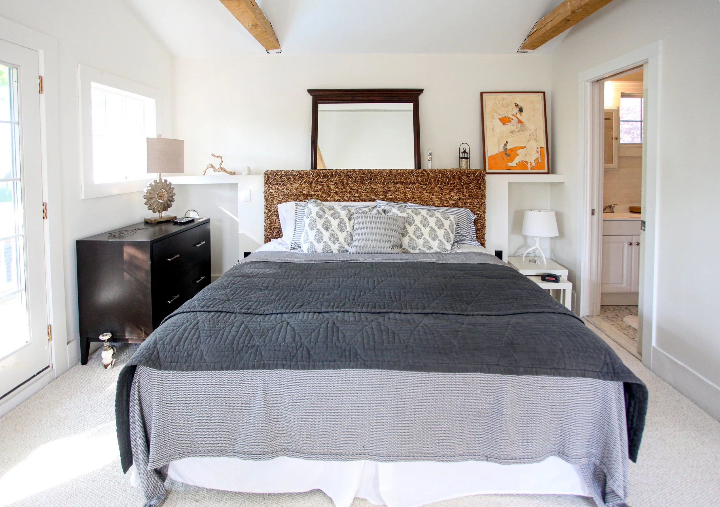 A cozy bedroom with a bed that has a woven headboard, gray bedding, and decorative pillows. There is a dark nightstand on the left side with a lamp, a decorative piece, and remote controls. A white nightstand with a lamp is on the right side. Two woo