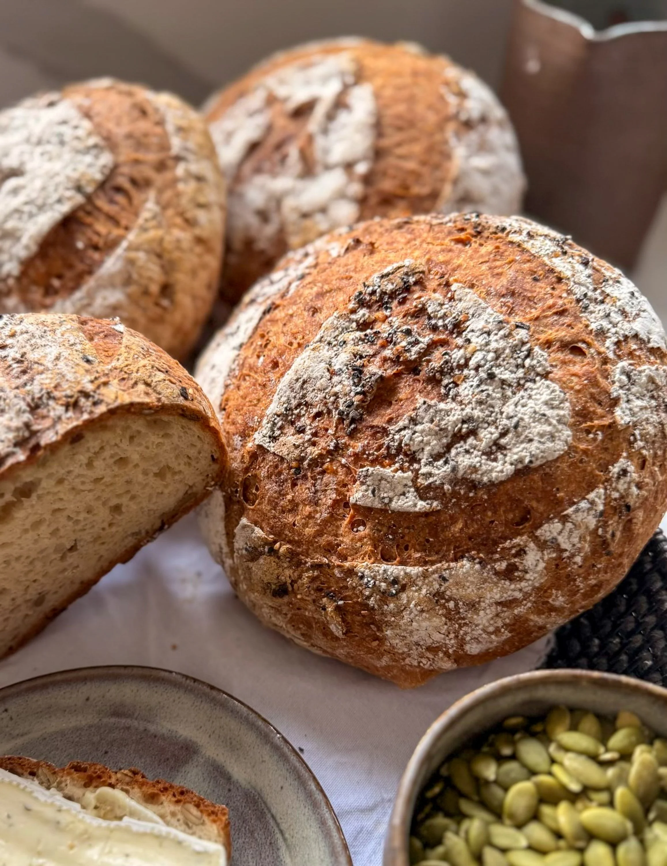 Close-up of three rustic round loaves of bread with powdered flour on top, positioned on a white cloth, accompanied by bowls of creamy dip and green pumpkin seeds.