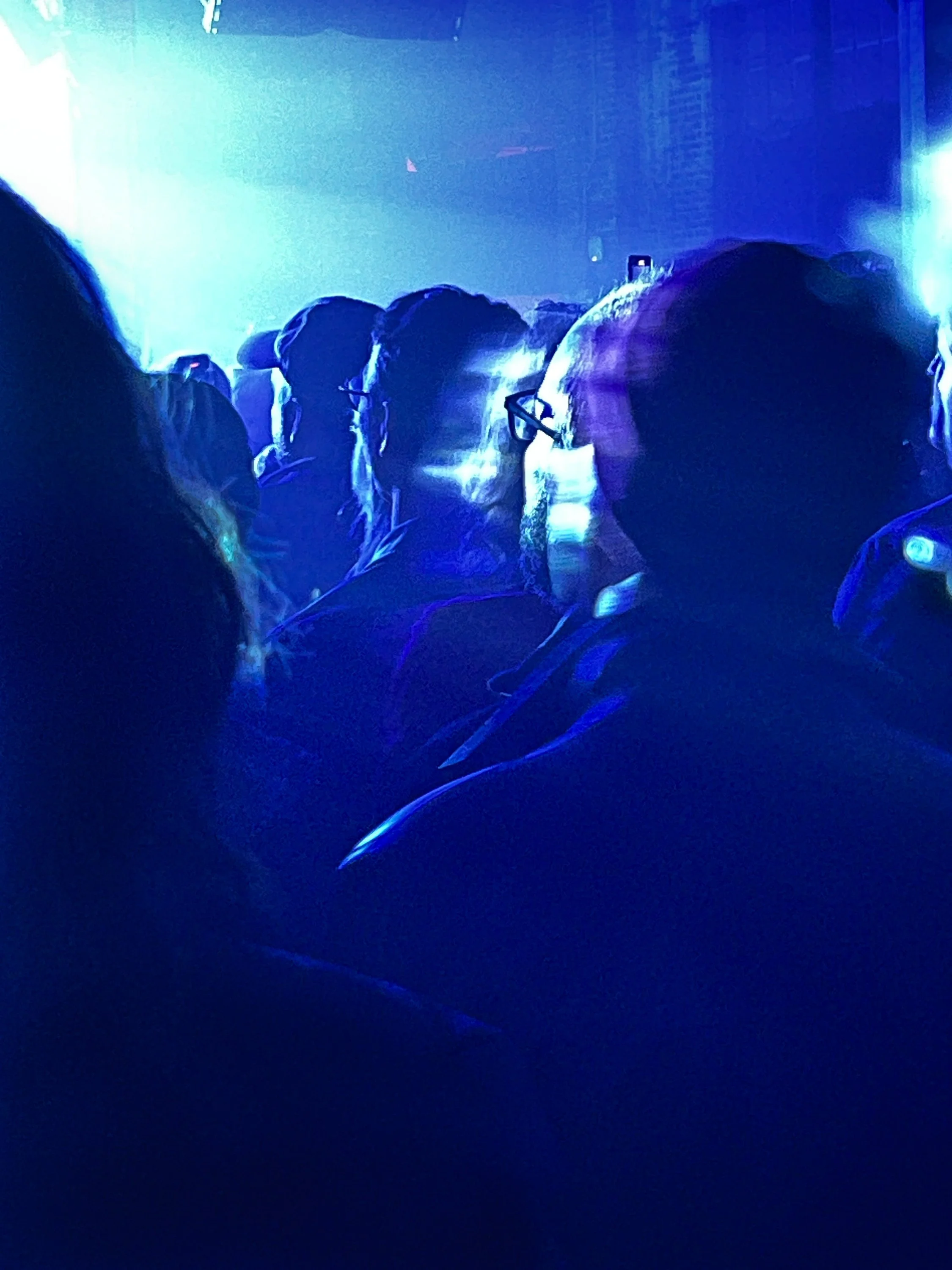 Audience members at a concert or event with blue lighting, some wearing glasses, and someone taking a photo or video with their phone.