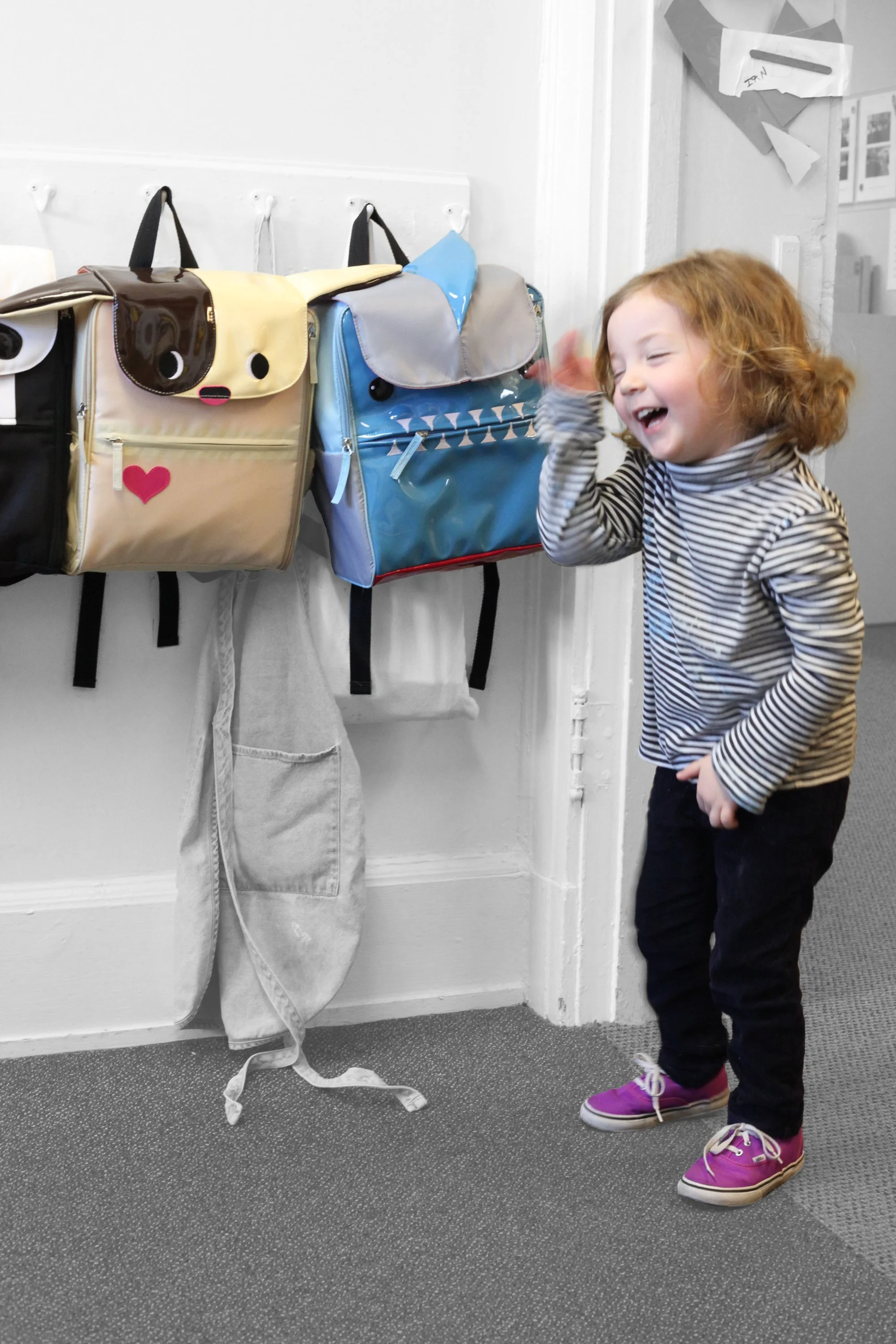 A young girl with curly red hair, wearing a striped long-sleeve shirt, black pants, and pink shoes, laughing and waving her hand near colorful backpacks hanging on hooks on a wall.