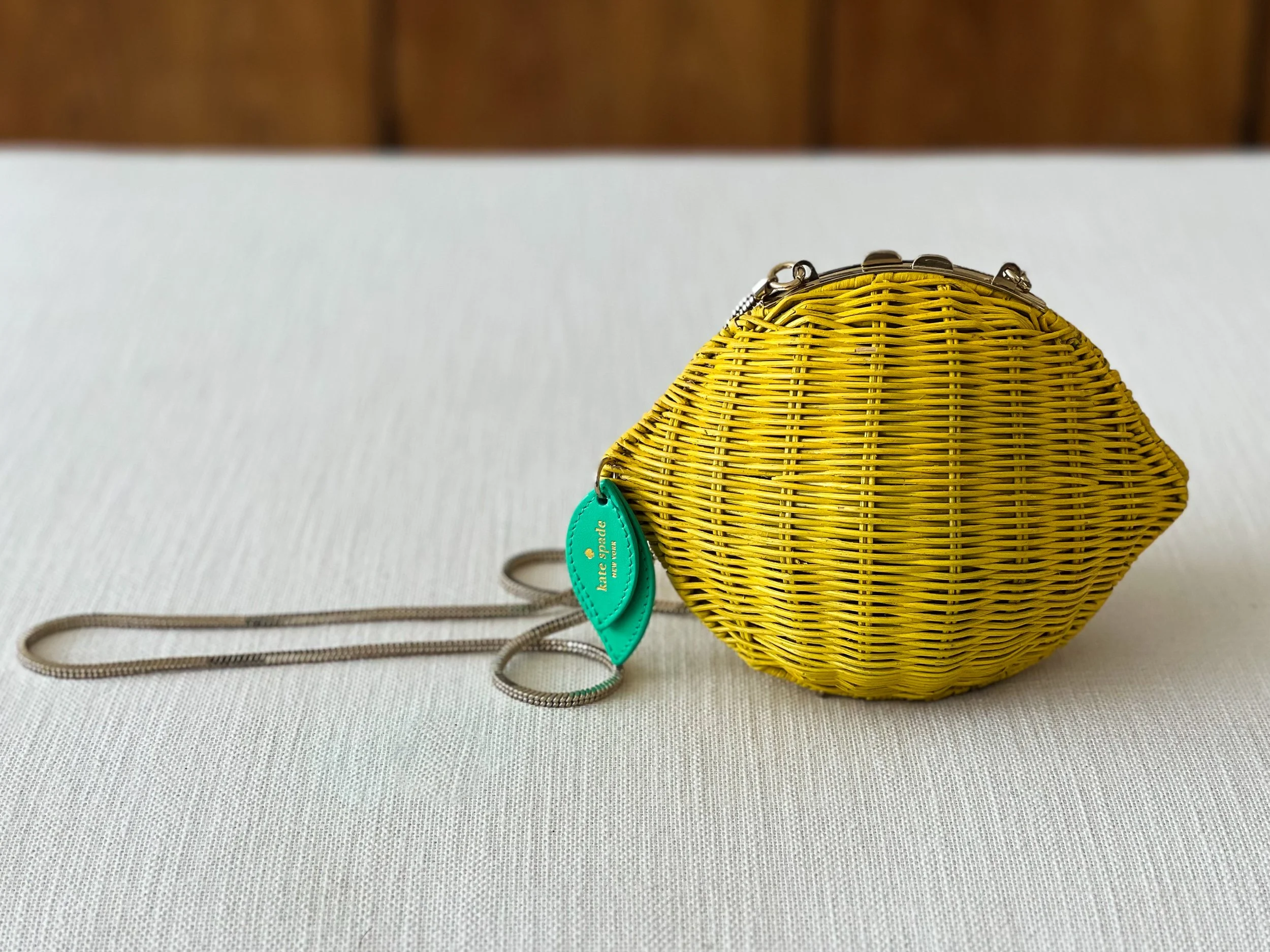 A yellow woven Kate Spade clutch handbag with a silver clasp, teal tags, and a beige strap on a light-colored textured surface.