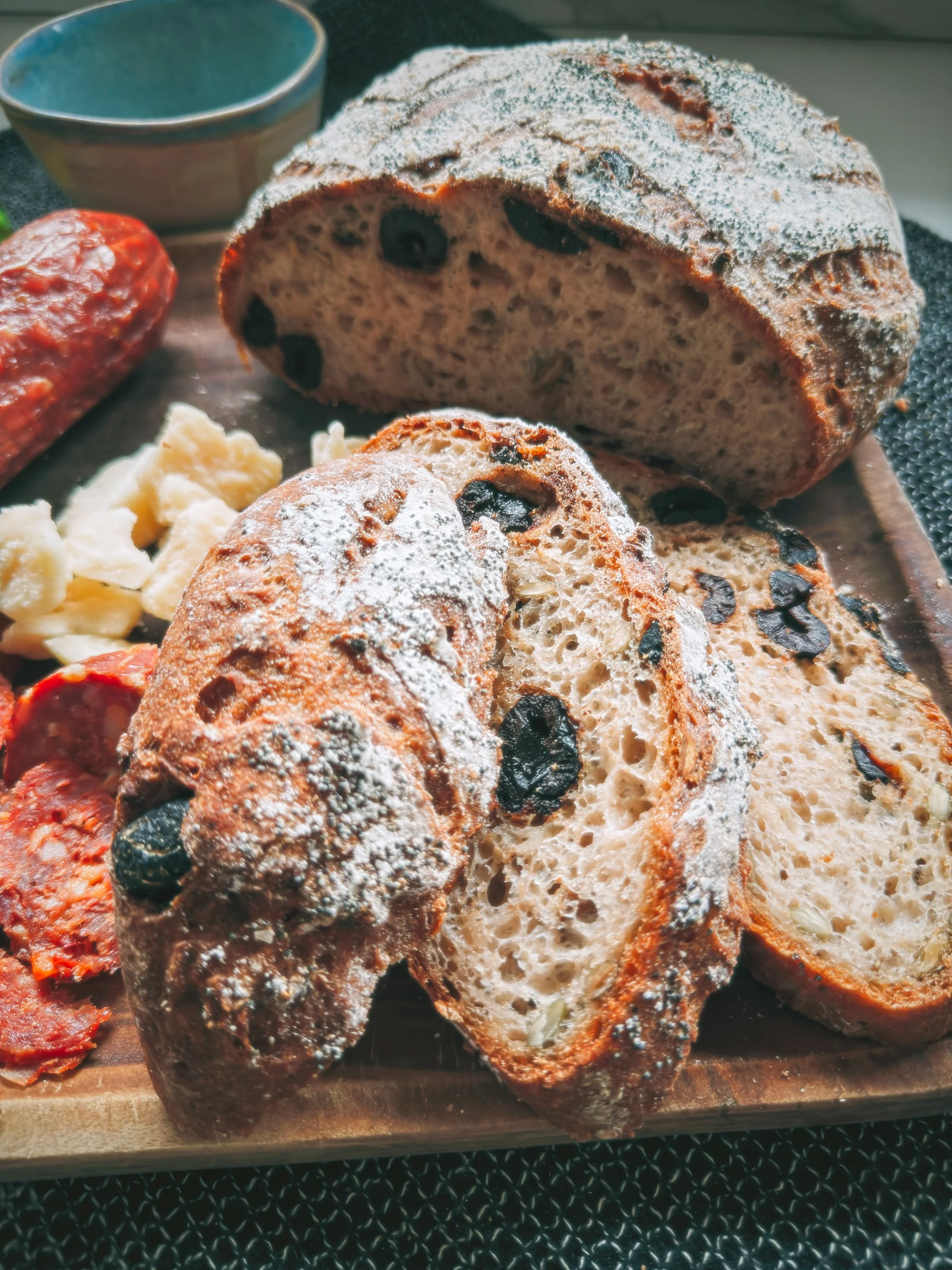 Sliced loaf of bread with black olives, dusted with flour, placed on a wooden cutting board. A piece of cooked sausage and some cheese are also visible, with a small bowl in the background.