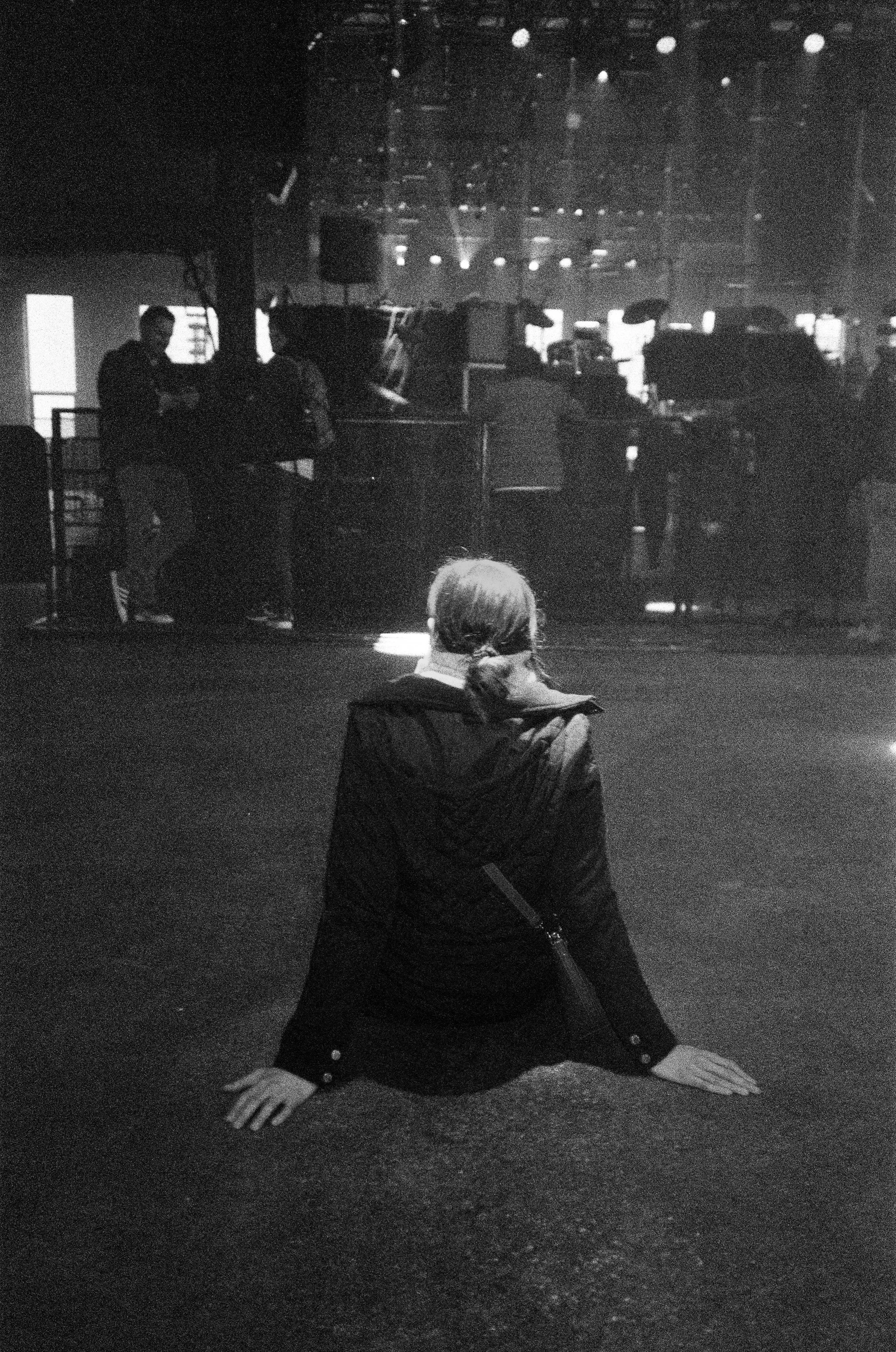 A woman in a dark jacket is sitting on the floor with her hands and arms supporting her, in a pose resembling a yoga or stretching position. In the background, there are several people standing and interacting near what appears to be a stage or perfo