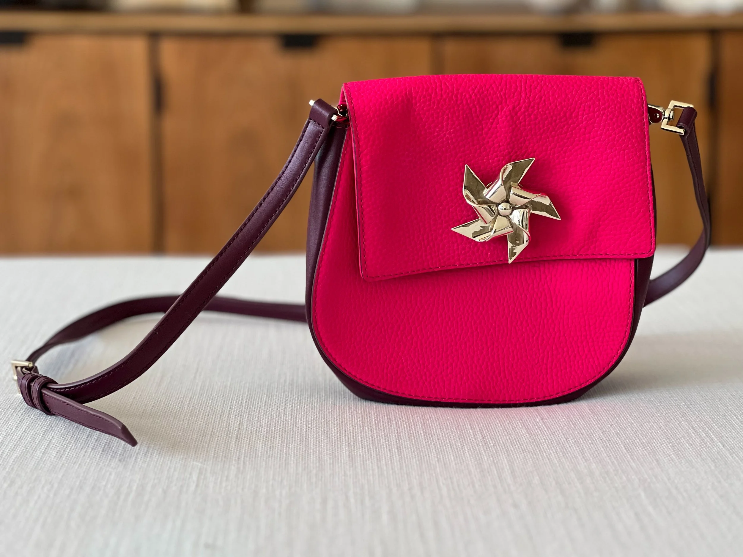 A pink Kate Spade handbag with a textured surface and a gold decorative clasp, resting on a fabric surface with a wooden backdrop.