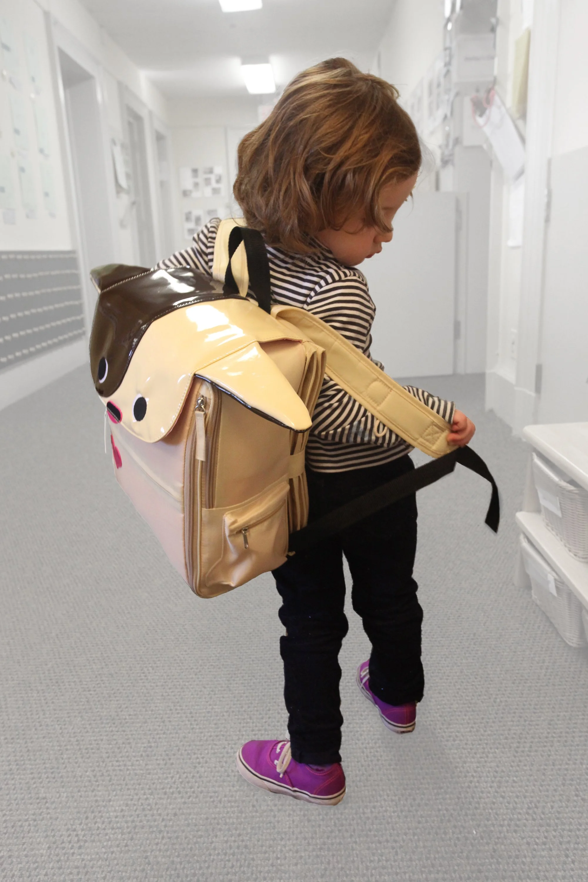 A young girl with short curly blonde hair wearing a striped long sleeve shirt, black pants, and purple sneakers. She is carrying a large beige backpack by Warm Milk, Inc. with a cat face design on the back and is looking down, standing in a hallway.