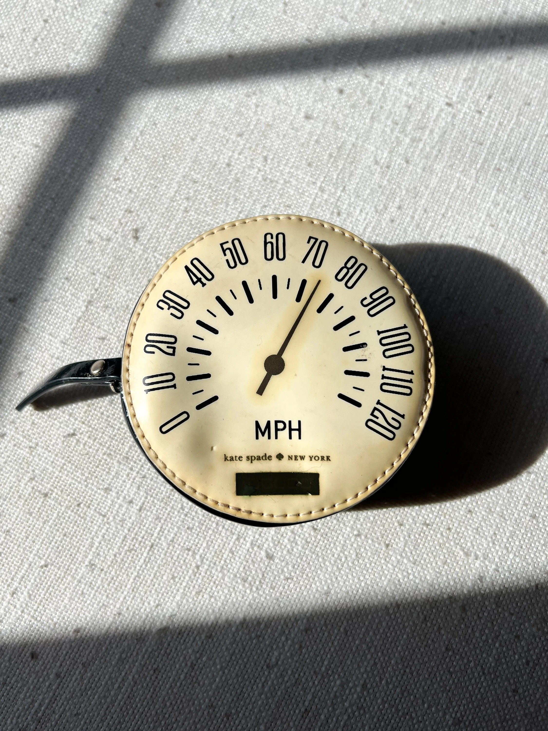 A vintage Kate Spade stopwatch with a cream face, black numerals, and black hands, displaying a speed of approximately 86 MPH, resting on a textured white fabric surface with shadows.