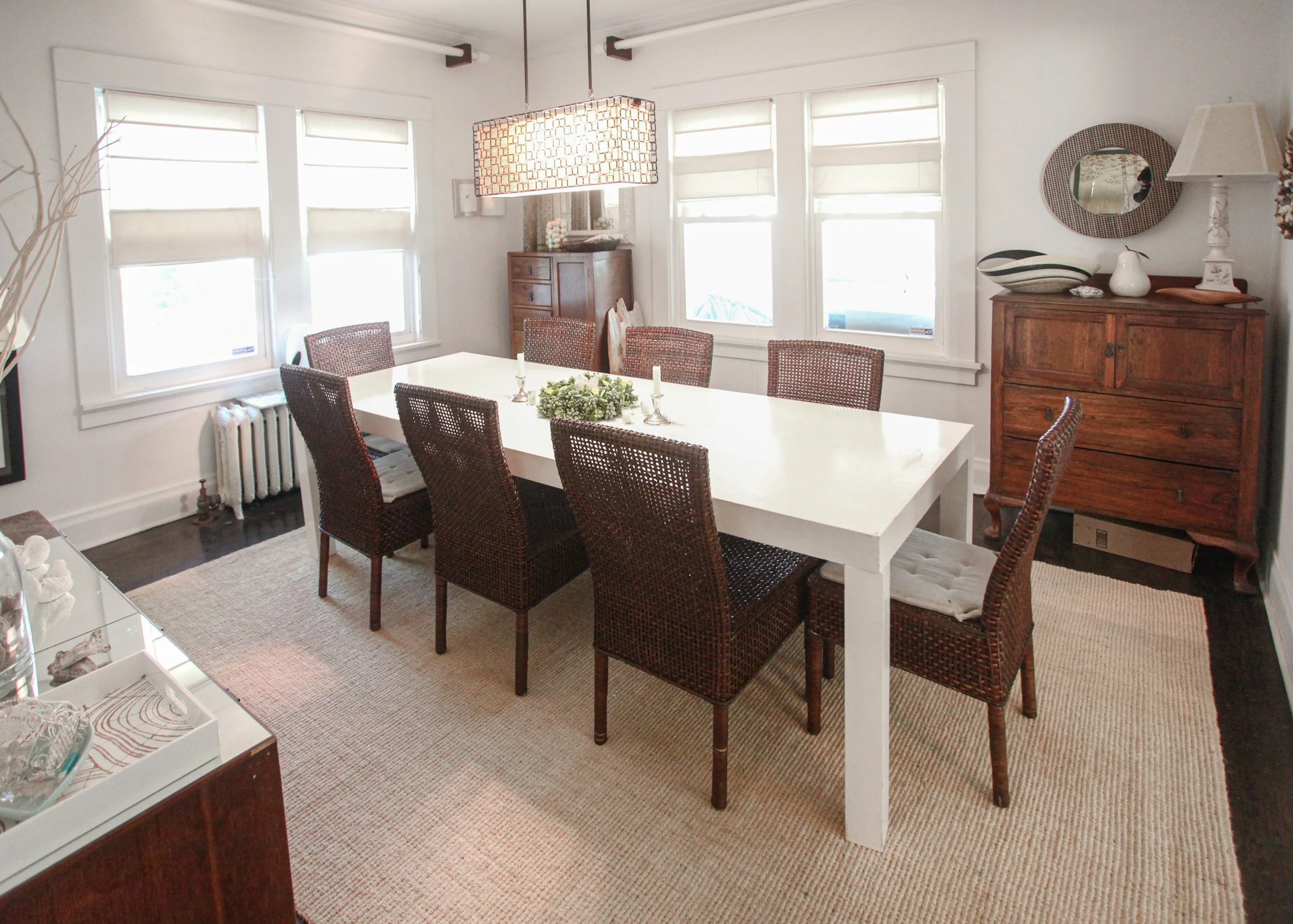 A dining room with a white table, six wicker chairs, three windows with white blinds, a wooden cabinet, and decorative items including plates, white pitcher, lamp, and mirror.