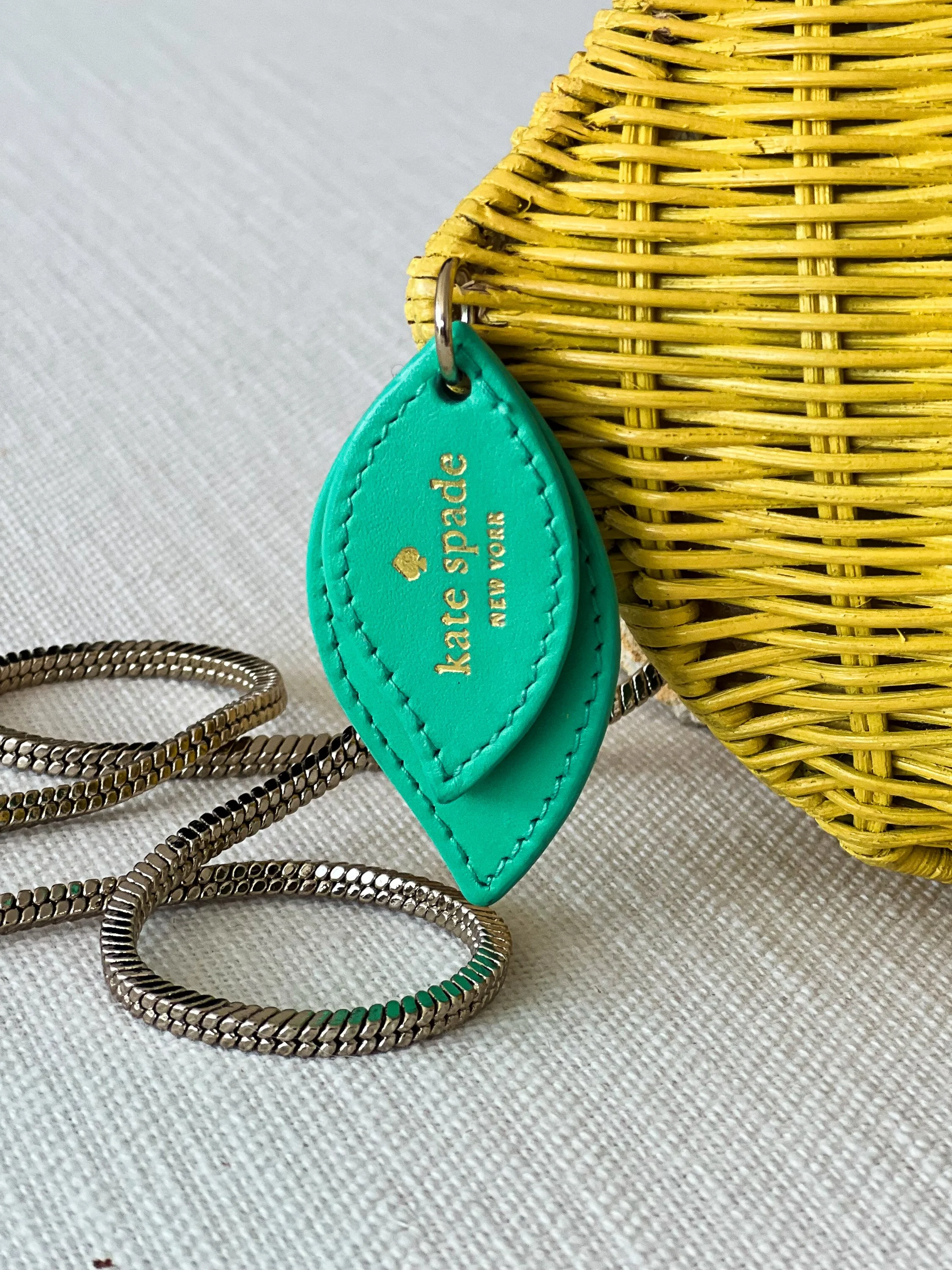 Close-up of a yellow woven Kate Spade clutch handbag, a turquoise leather name tag with gold lettering, and silver jewelry on a white fabric surface.