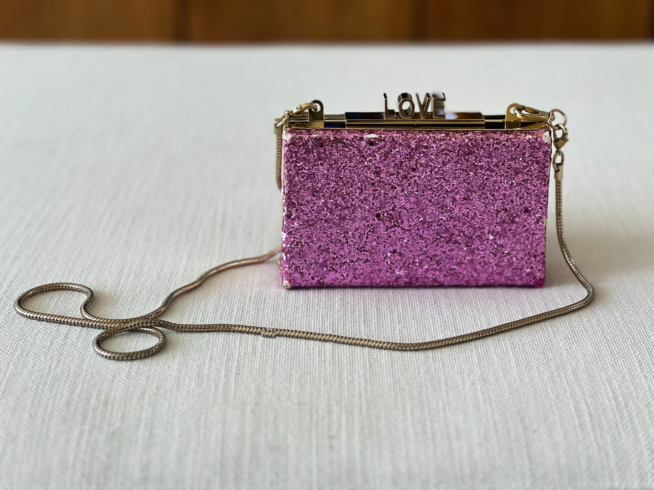 A small, rectangular, pink glittery purse with a gold chain strap and a decorative gold butler clasp that spells out the word 'LOVE' on top.