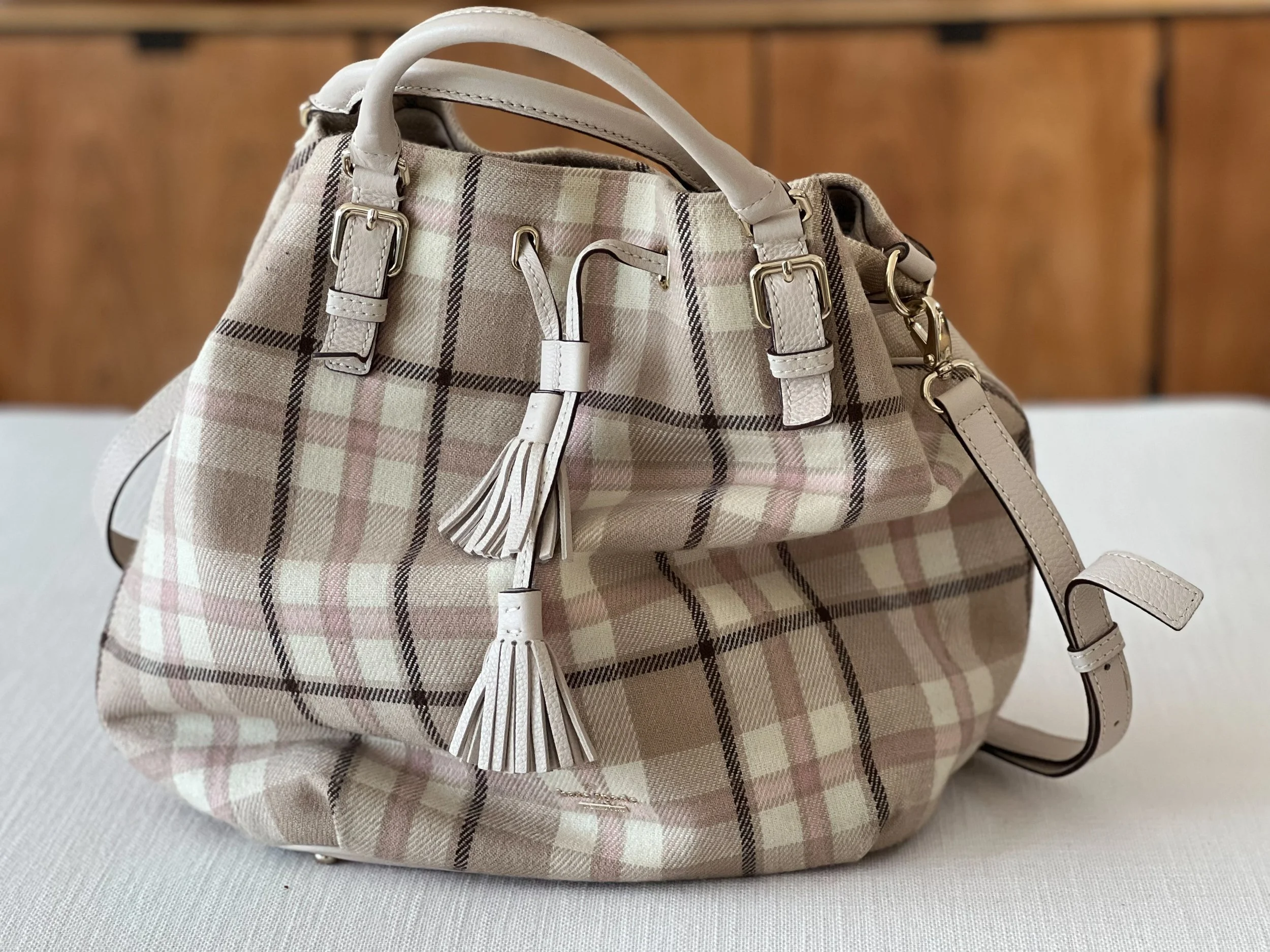 A beige Kate Spade handbag with a plaid pattern, beige leather straps, and gold hardware, placed on a light-colored surface with a wooden backdrop.