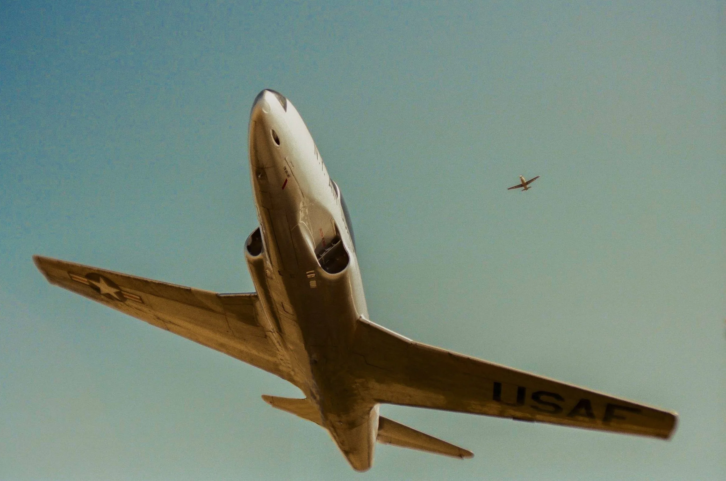 Military aircraft flying with another smaller plane in the background against a blue sky.