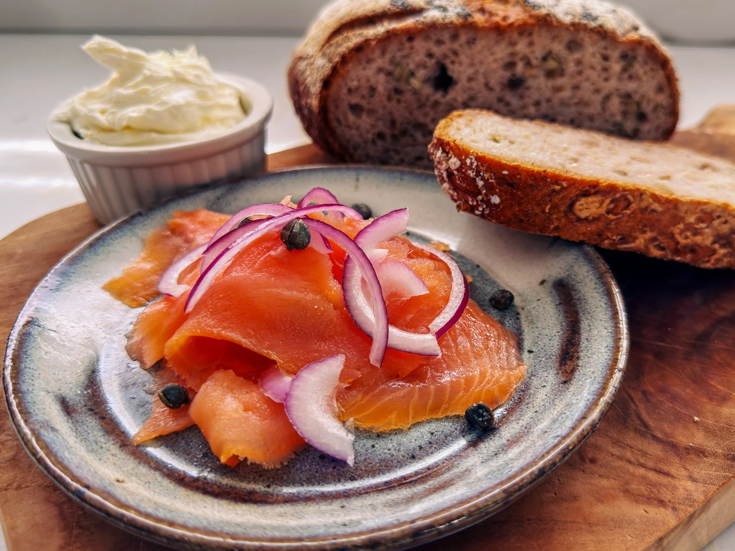 Slices of smoked salmon with thinly sliced red onions and capers on a ceramic plate, with a side of smoked salmon on a separate plate, topped with chopped herbs, and a bowl of sour cream in the background, all on a wooden surface.
