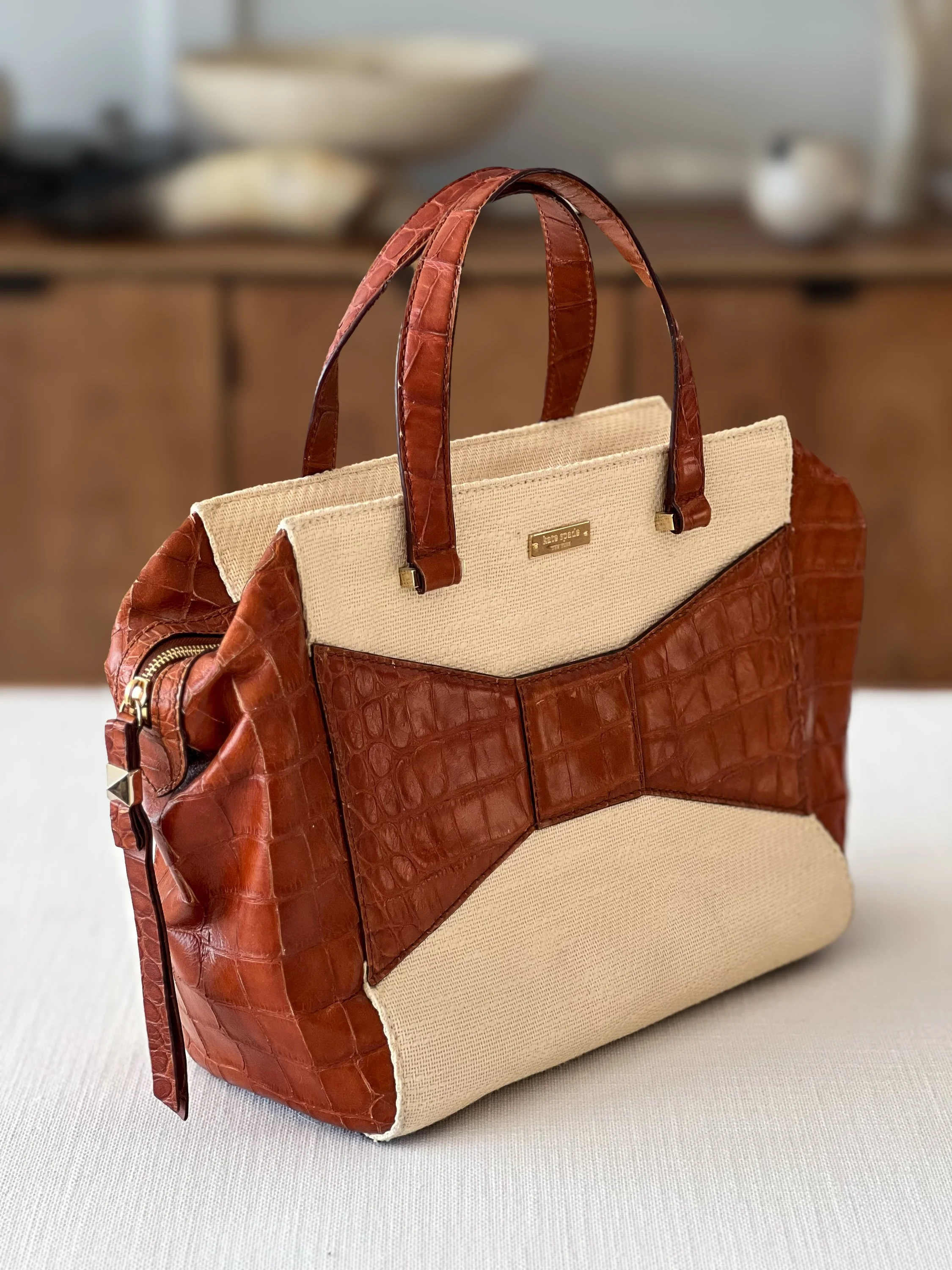 A stylish Kate Spade tote bag with red croc-embossed leather accents and beige canvas body, placed on a neutral surface in a room with blurred background.