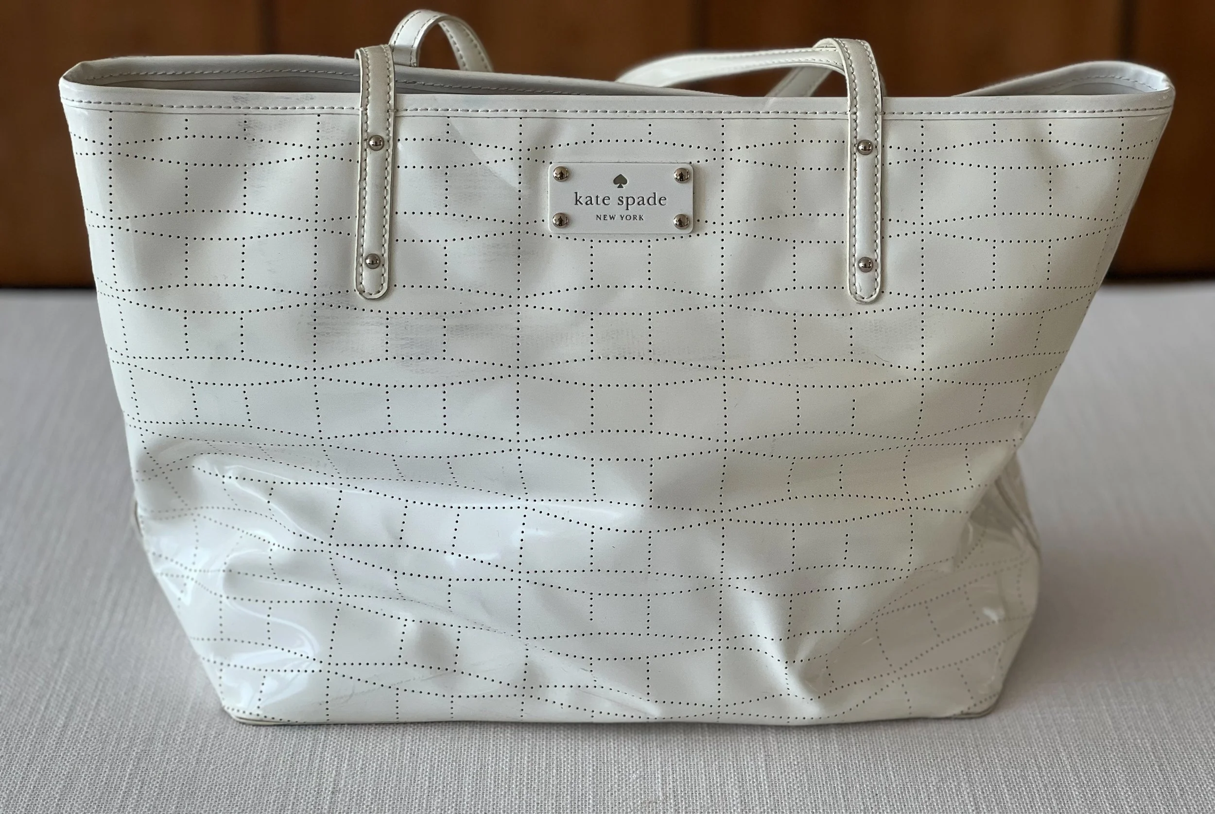A white Kate Spade tote bag with a perforated dotted pattern and silver stud accents, featuring a label with the brand "Kate Spade New York".