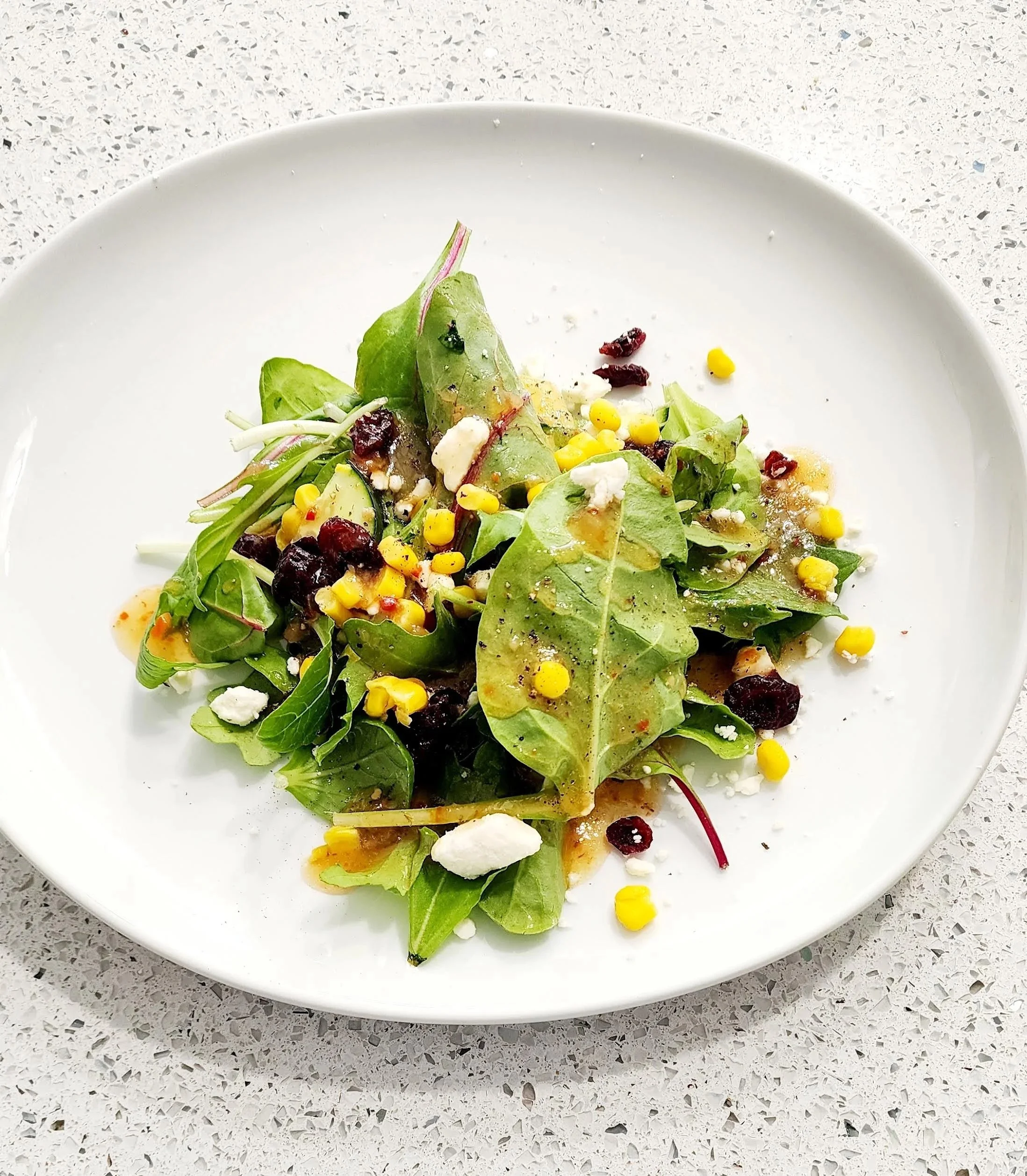 Mixed green salad with corn, cranberries, cheese, and dressing on a white plate.