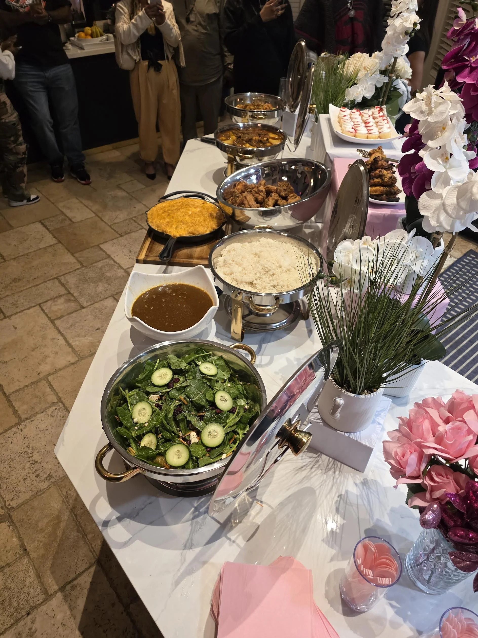 A buffet table with various dishes including rice, salad, gravy, fried chicken, and desserts. Pink and white flowers decorate the table, with people standing around in a room with stone tile flooring.