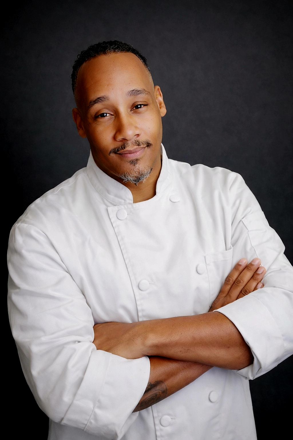 Portrait of a male chef wearing a white chef's coat with arms crossed, standing against a dark background.