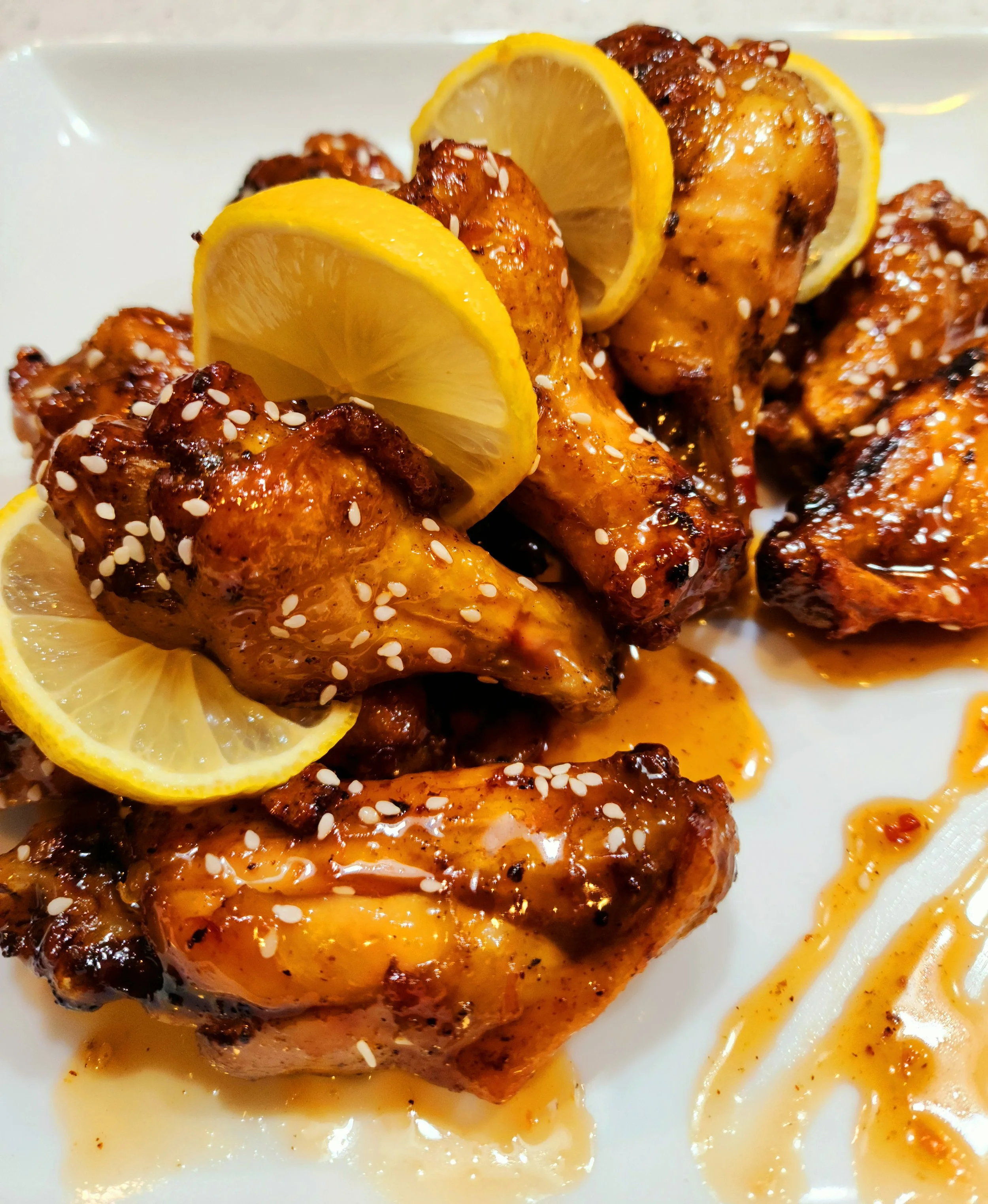 Close-up of three grilled chicken drumsticks with lemon slices and sesame seeds on a white plate.