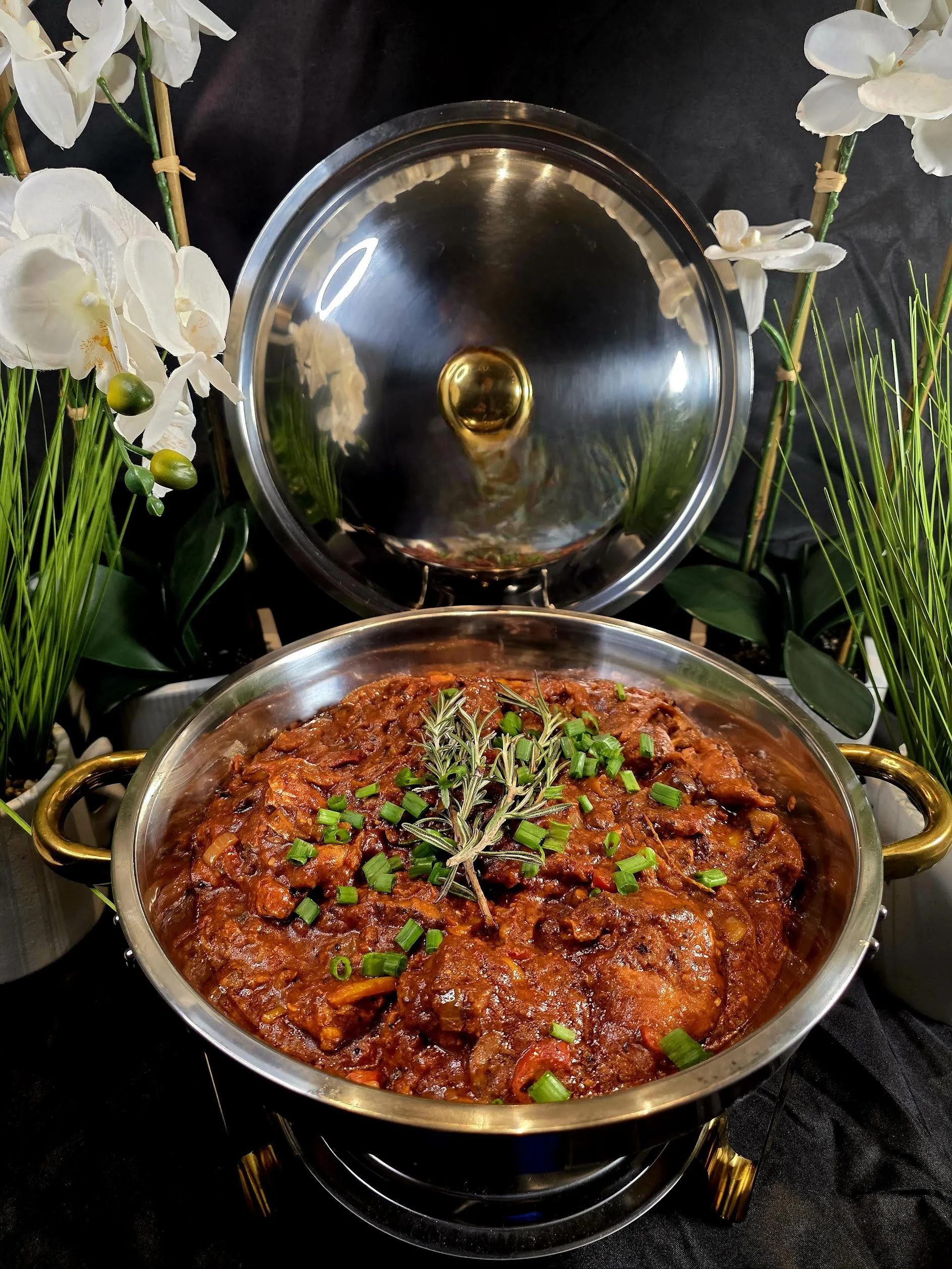 A stainless steel serving dish with a lid, filled with a beef stew garnished with chopped green onions and rosemary. The dish is surrounded by white orchids and green grass plants.