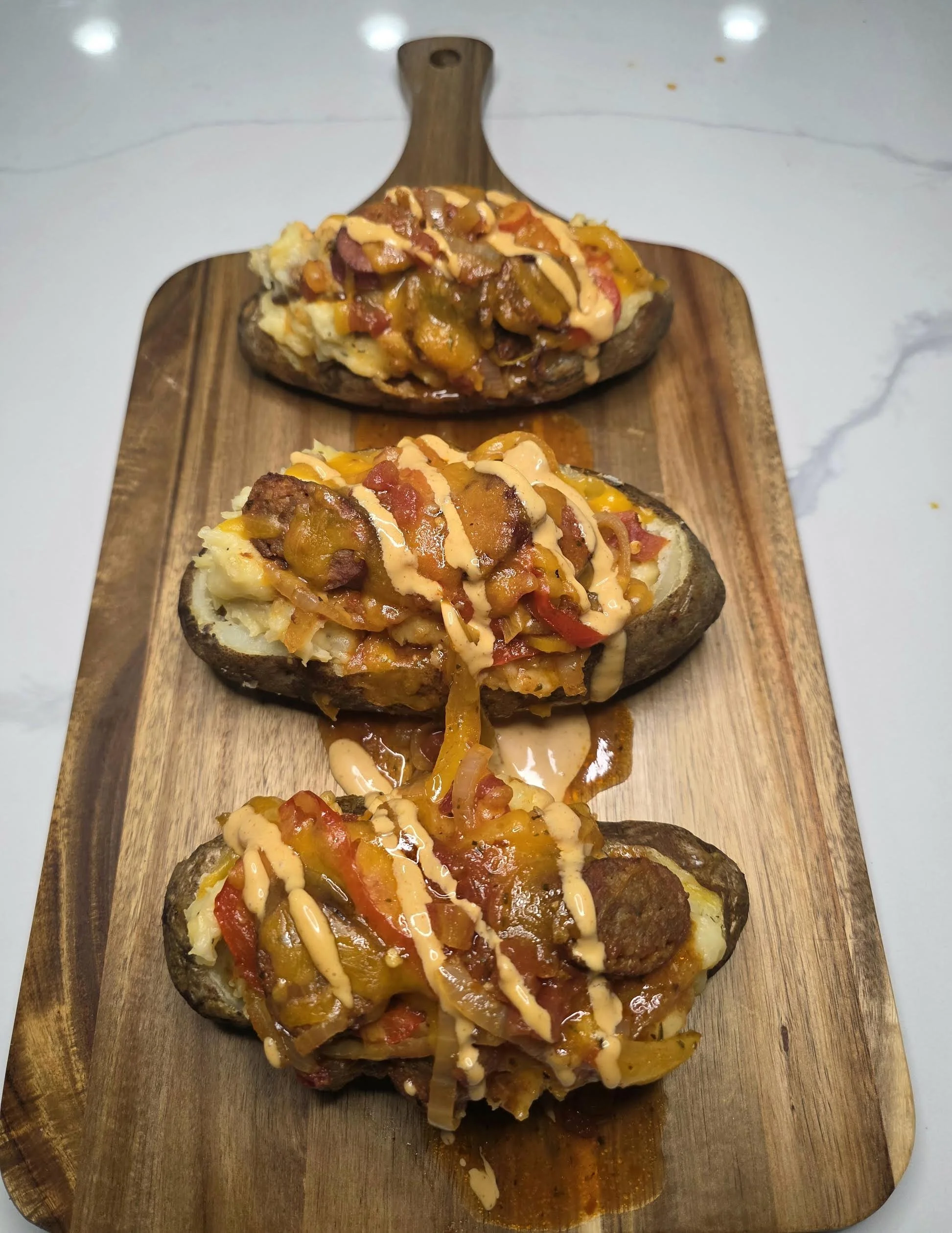 Baked potatoes topped with sausage and peppers and drizzle of sauce on a wooden serving board.
