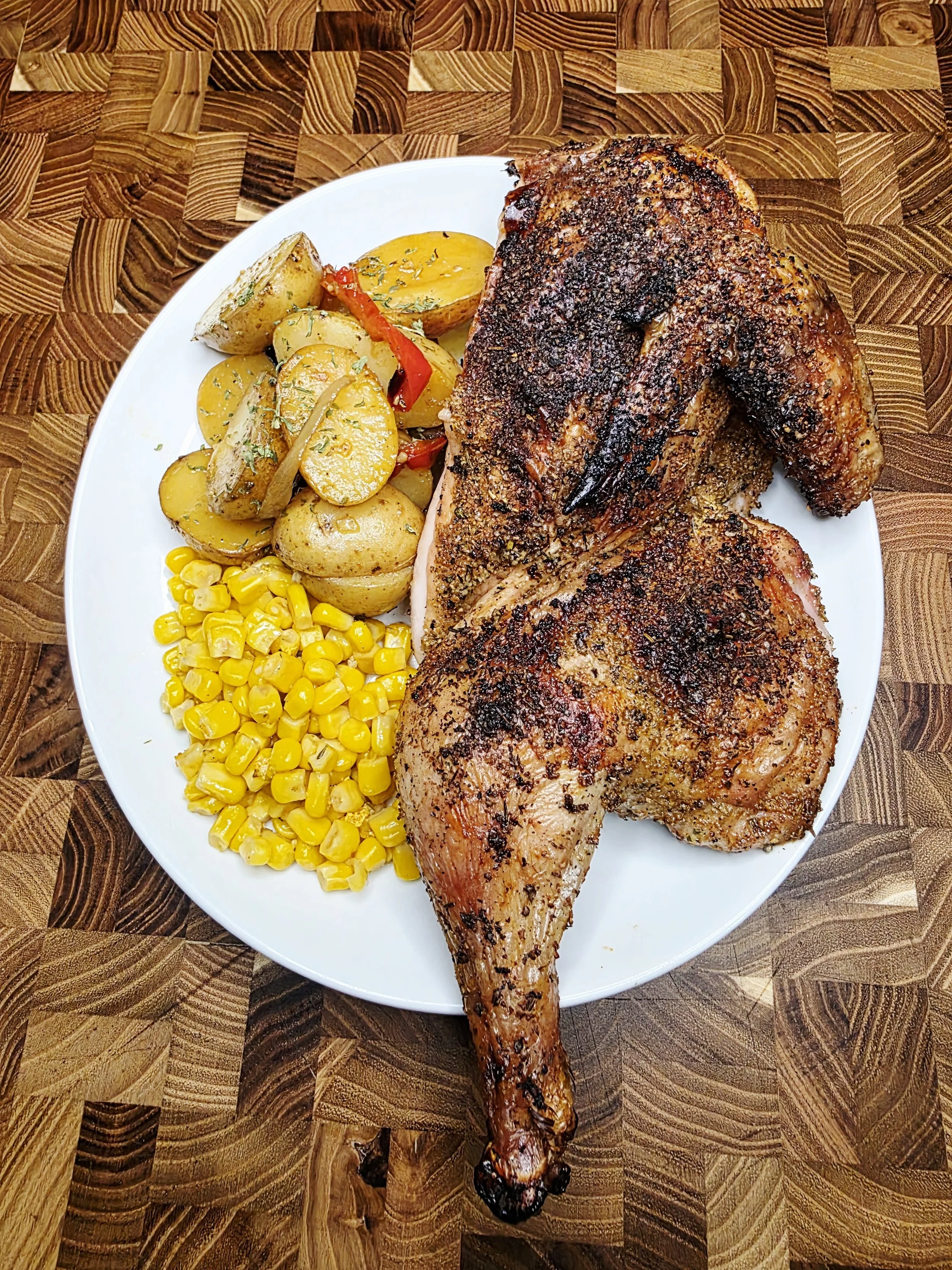 Roasted chicken leg quarter with seasoned crispy skin, accompanied by roasted potatoes with herbs, sliced bell peppers, and yellow corn kernels on a white plate.