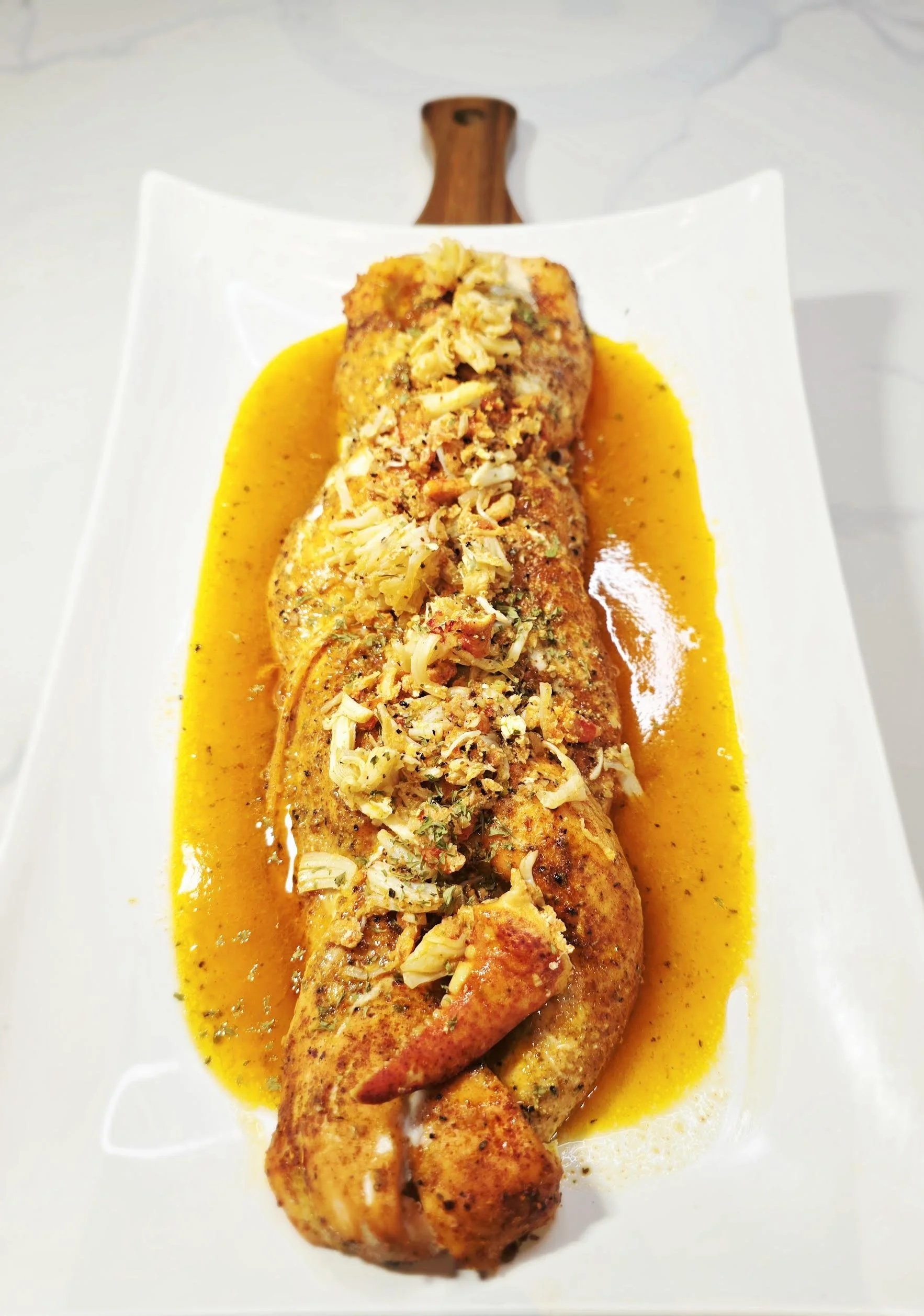 A cooked chicken breast topped with shredded cheese and herbs, served with a yellow sauce on a white rectangular plate with a wooden handle at the top.